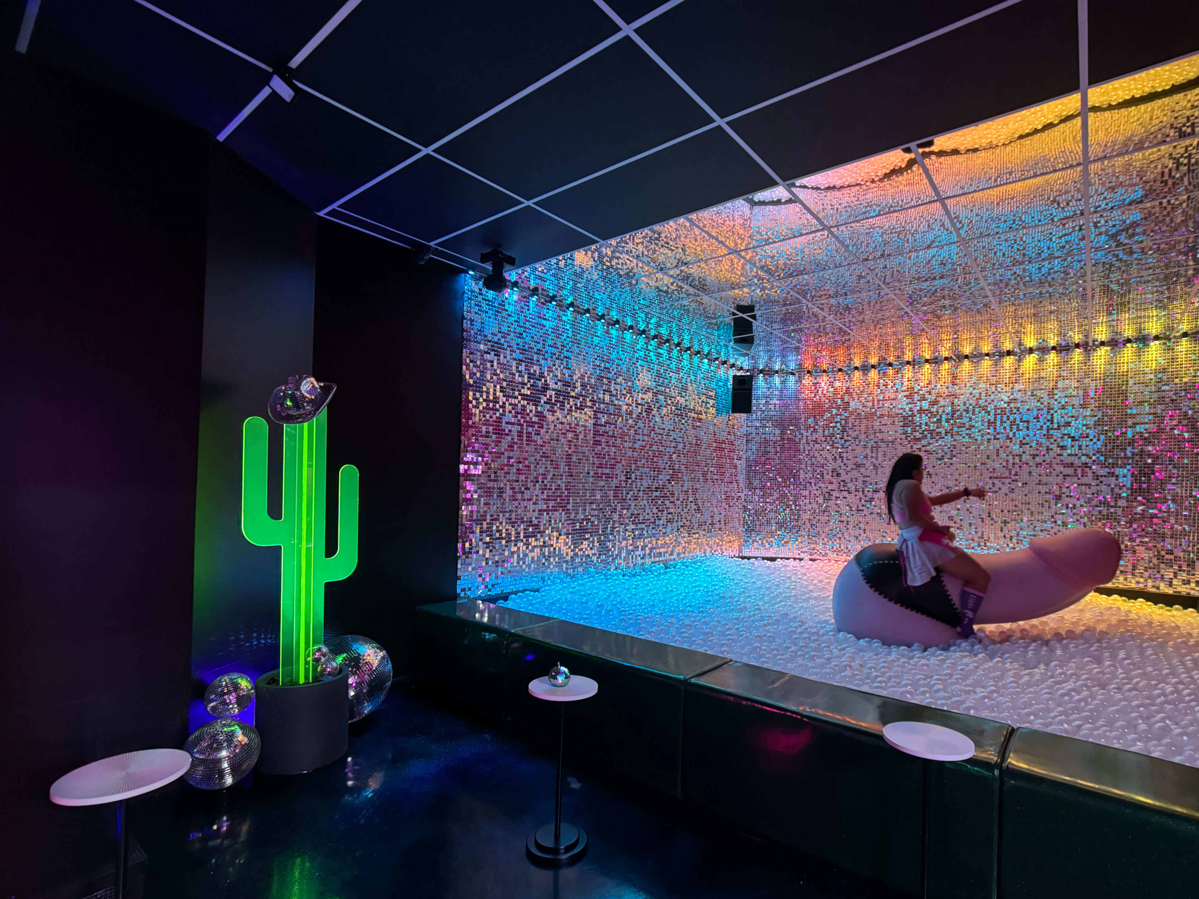 The scene features a colorful room with a mirrored wall, a large inflatable figure on a pool of white foam, a cactus-shaped light, and disco balls on small tables.