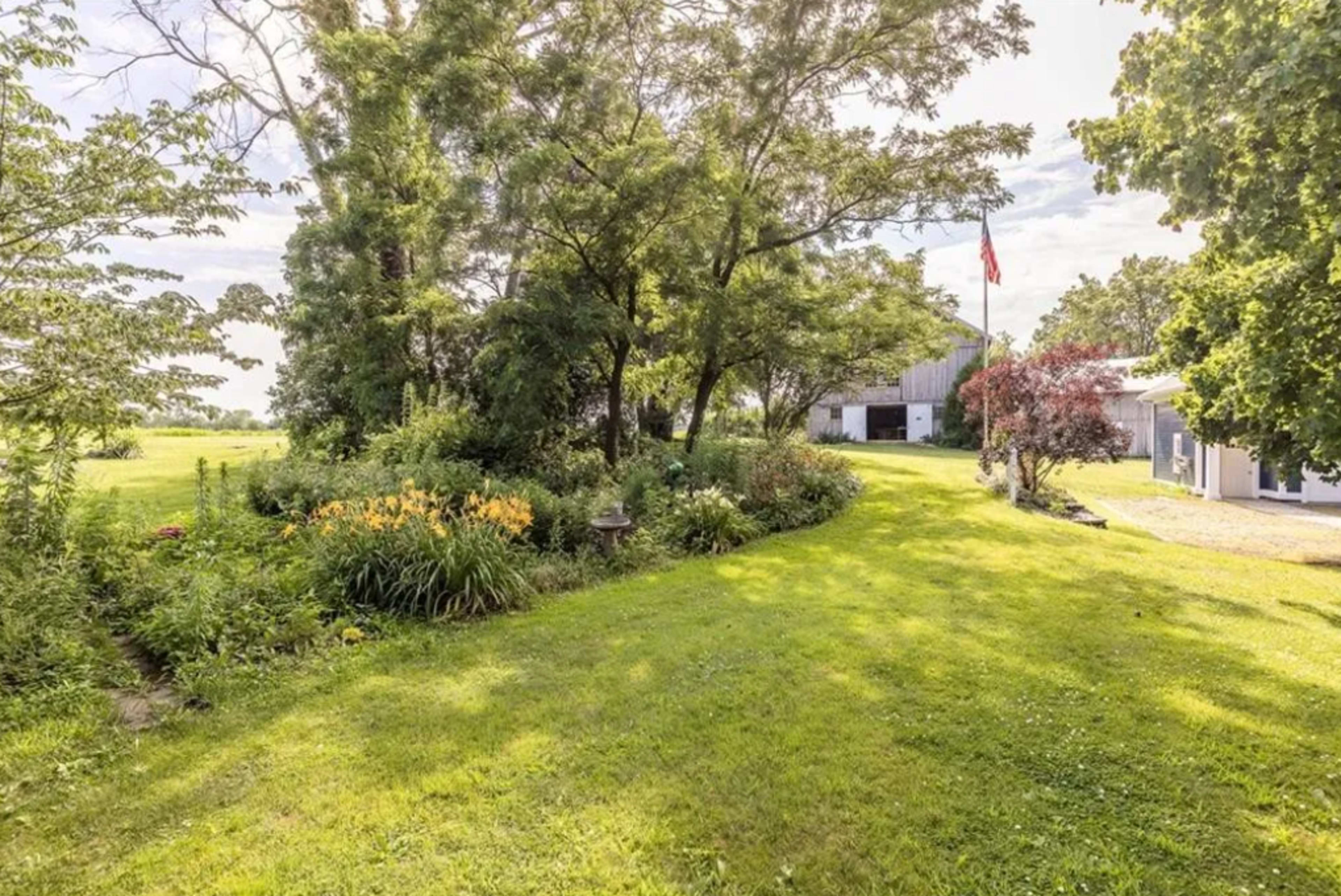 The scene features a grassy yard bordered by flowering plants and trees, leading to a building in the background, with an American flag visible among the foliage.