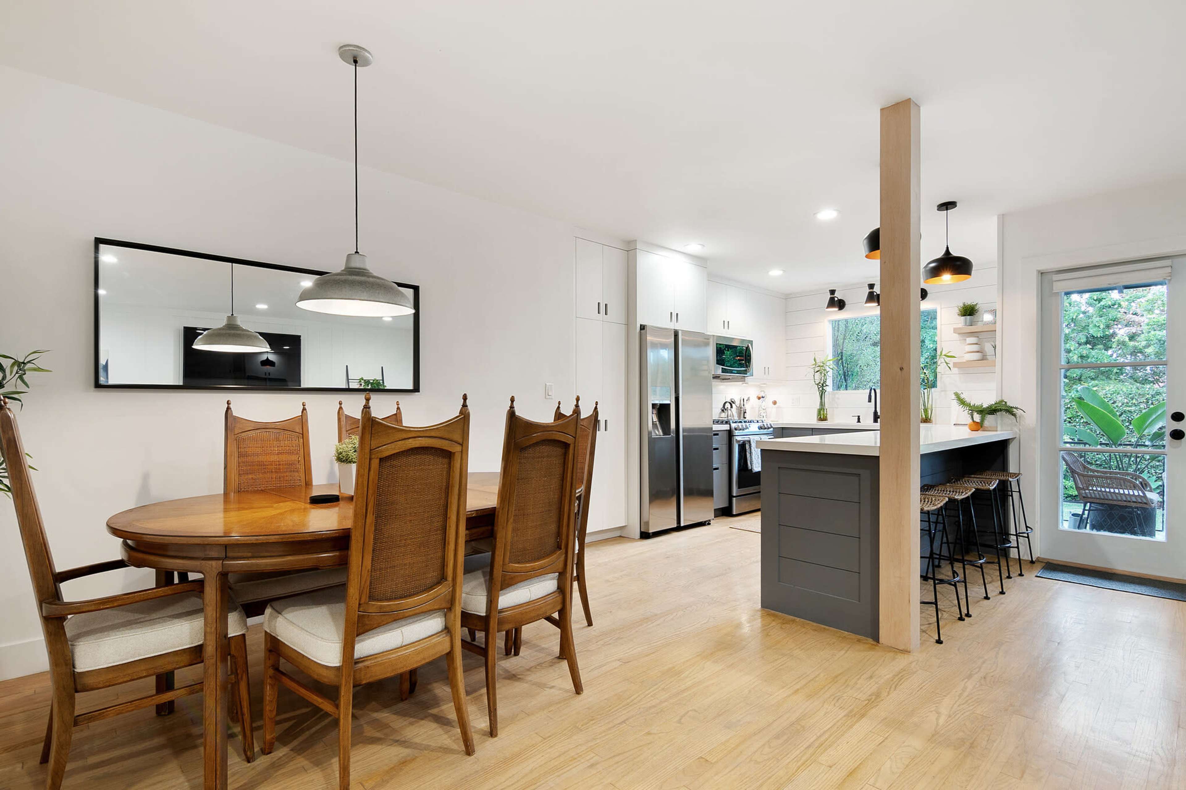 The image shows a modern kitchen and dining area with a wooden dining table, high-backed chairs, and sleek appliances.