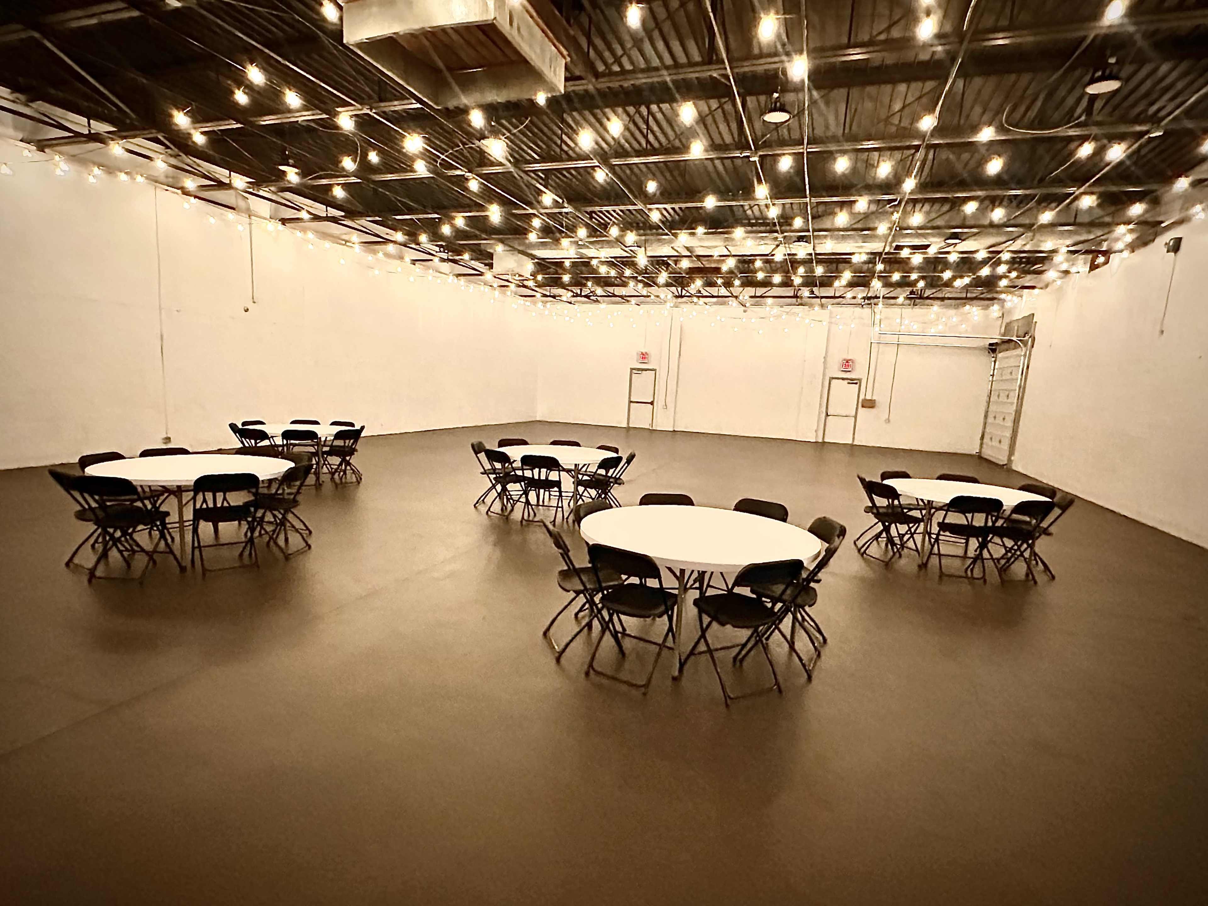 The image shows a large, empty indoor space with multiple circular tables and black chairs, illuminated by string lights above.