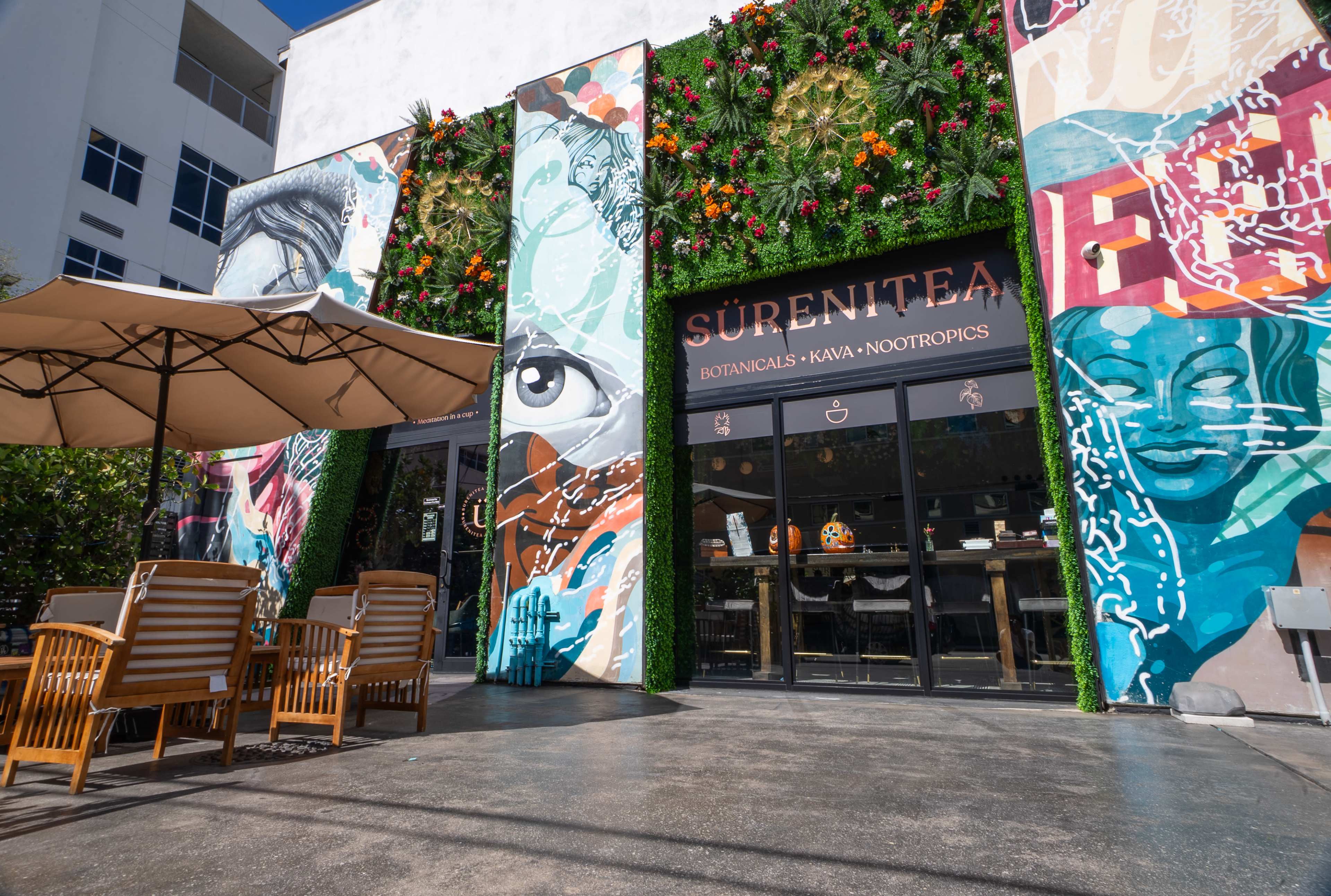 The image shows the exterior of a vibrant café named "Surenitea," featuring colorful murals and an outdoor seating area with wooden chairs under a large umbrella.