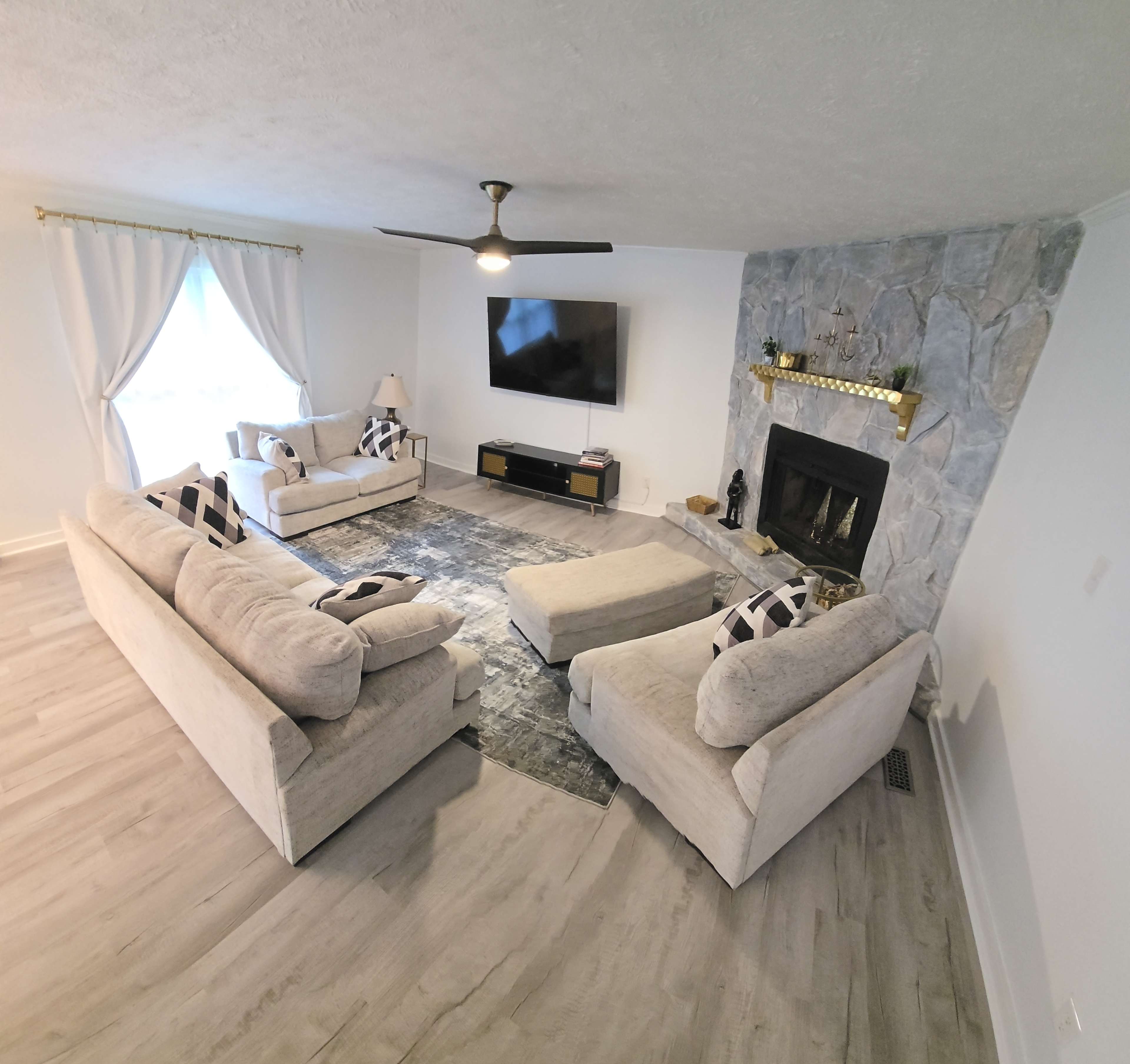 The image shows a spacious living room with two sofas and two armchairs arranged around a coffee table, featuring a stone fireplace and a television mounted on the wall.