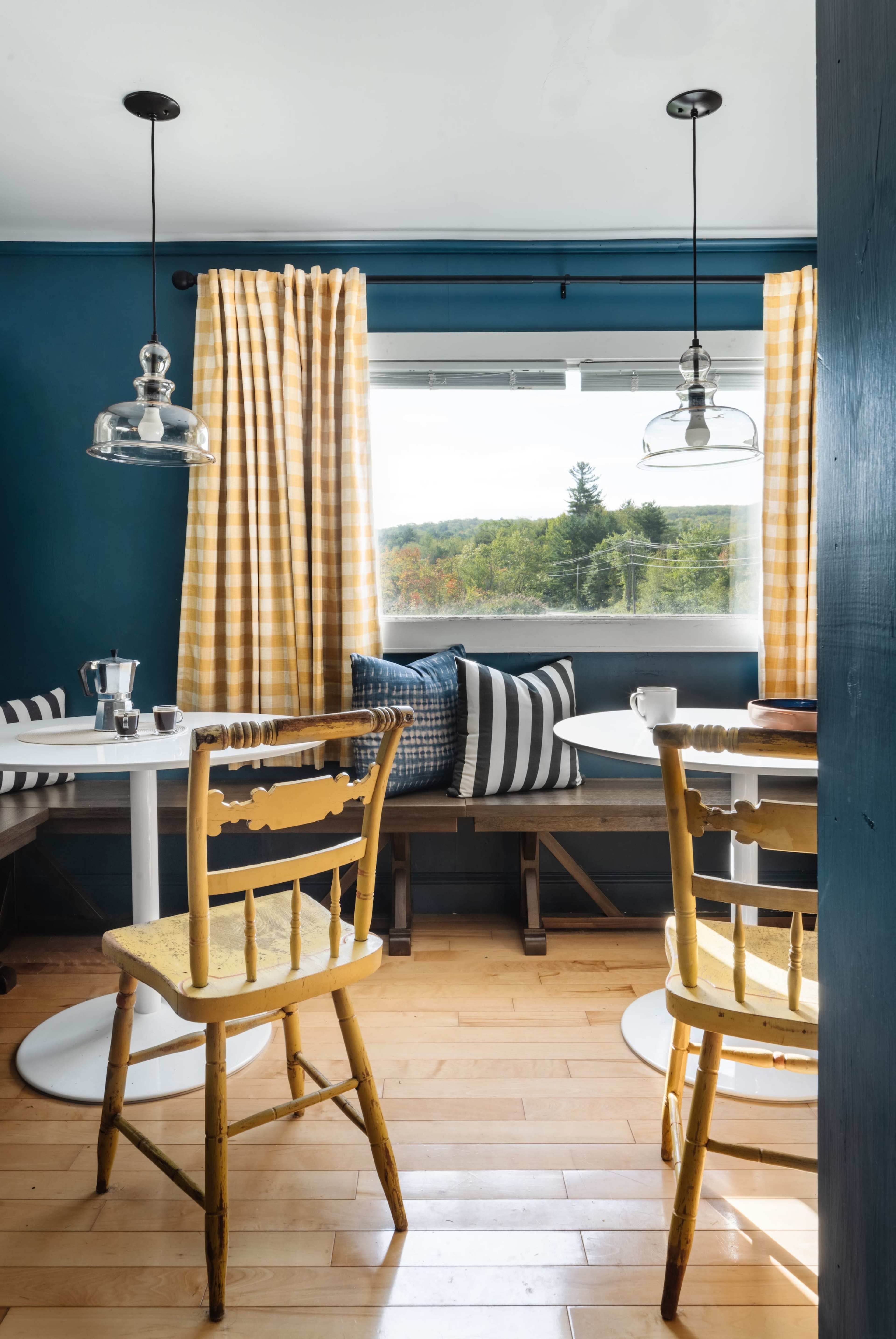A cozy dining area features a round table with yellow and white striped curtains, two hanging lamps, and a bench with striped cushions overlooking a scenic view.