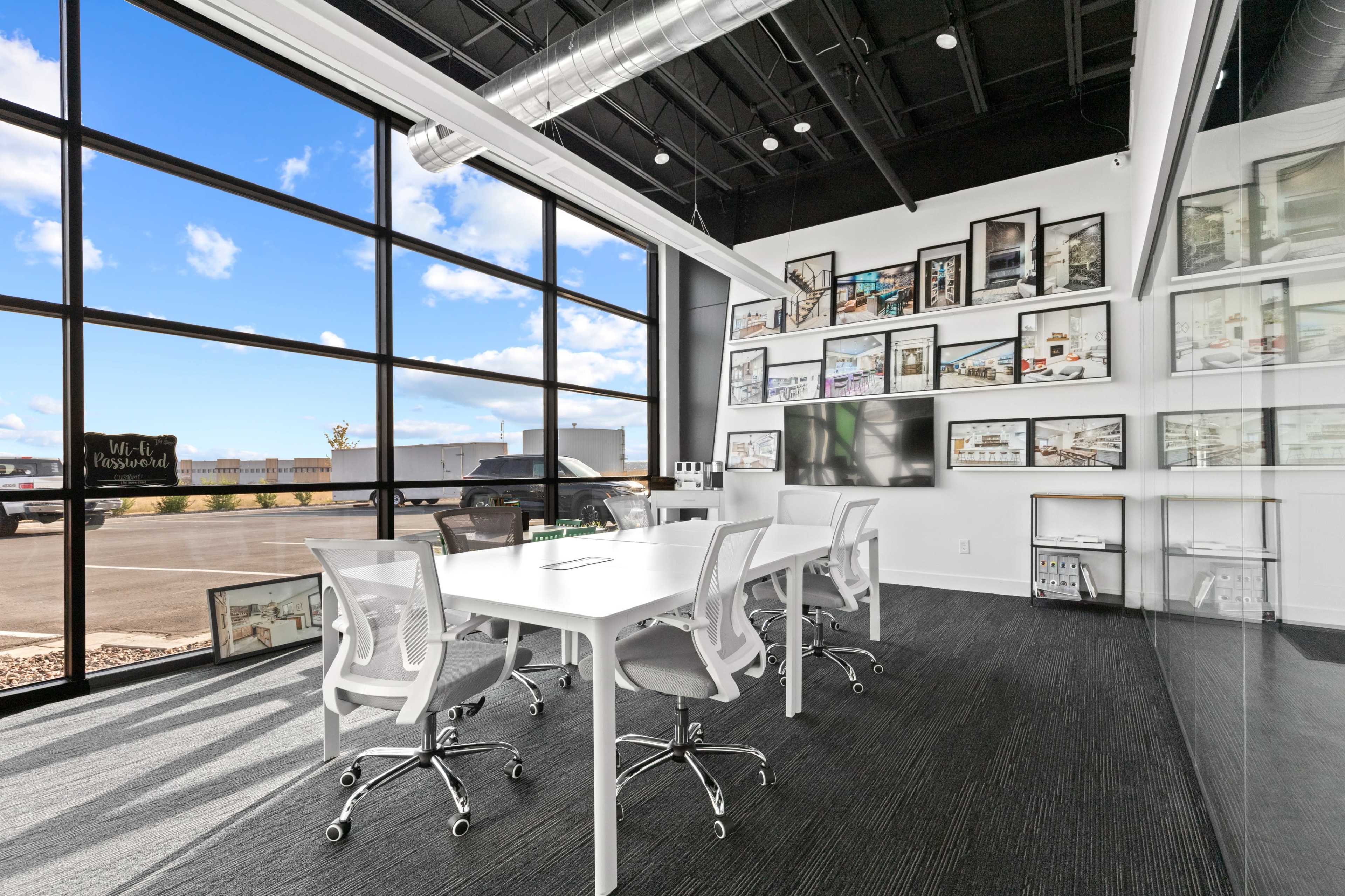 A modern conference room features a large glass wall with multiple framed images, a white table surrounded by chairs, and a sleek display screen.