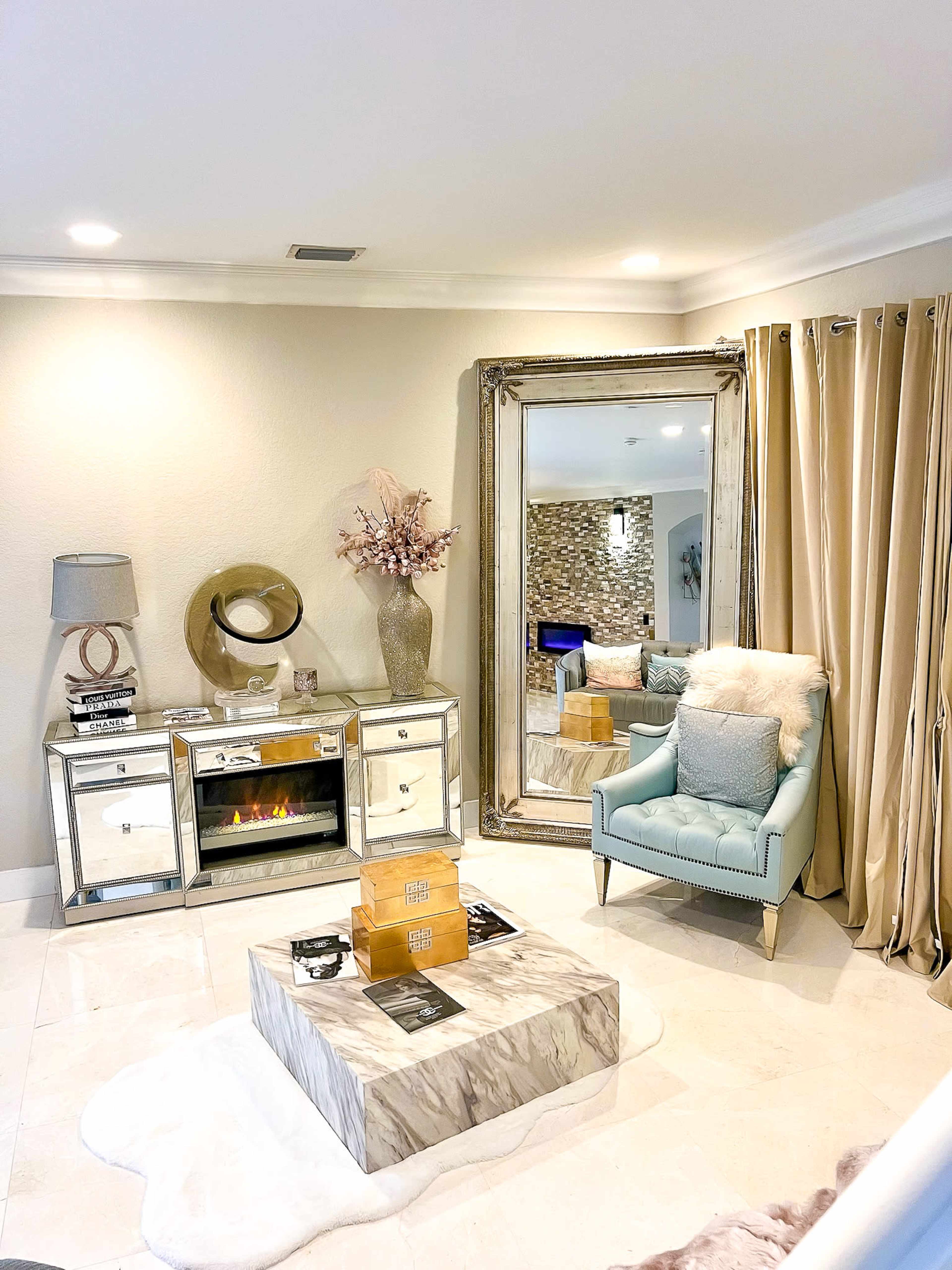 A modern living room features a light-colored decor, a large mirror, a blue chair, and a marble coffee table, with a decorative fireplace and curtains.