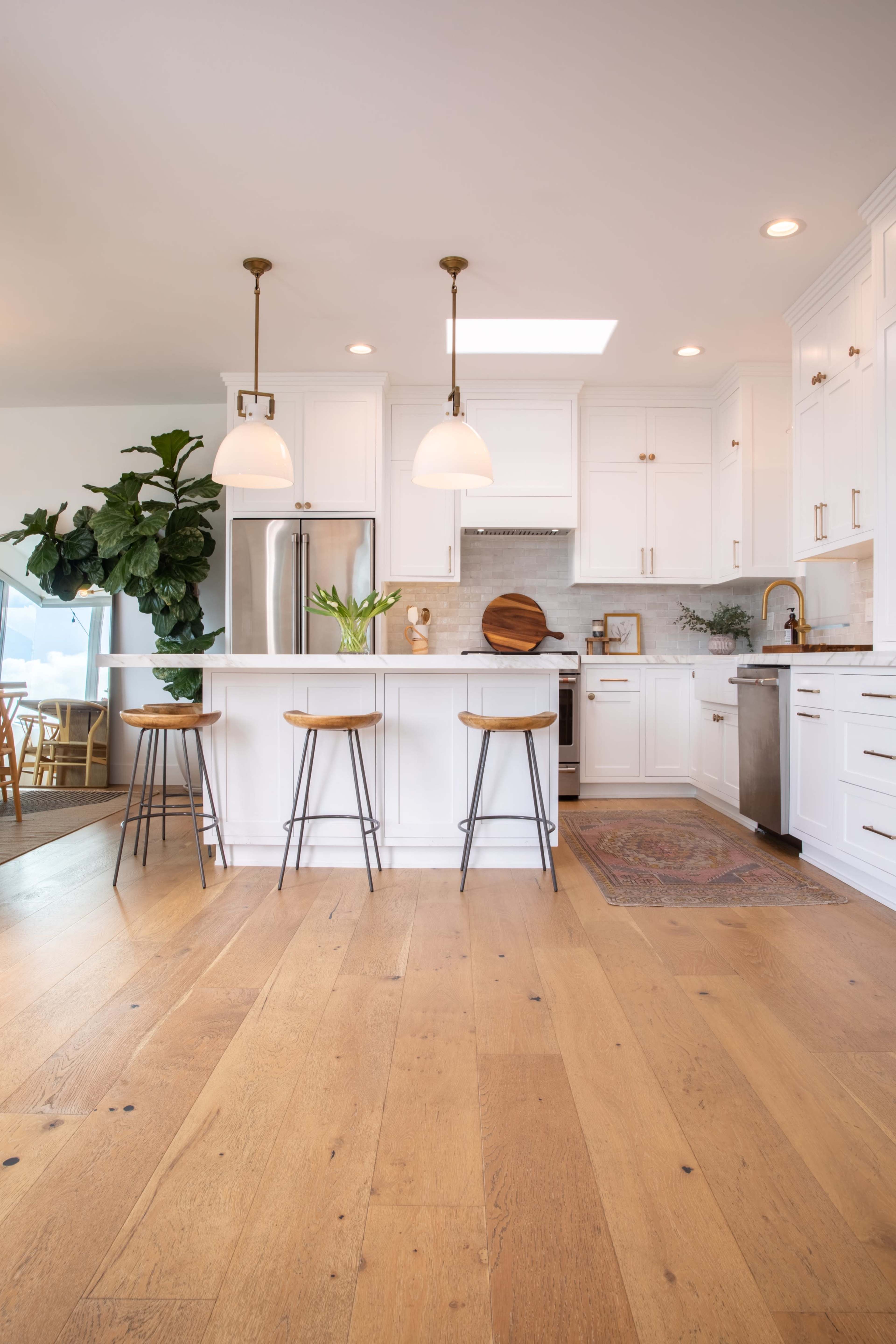 A modern kitchen featuring white cabinetry, a central island with three bar stools, and large windows that allow natural light to enter.