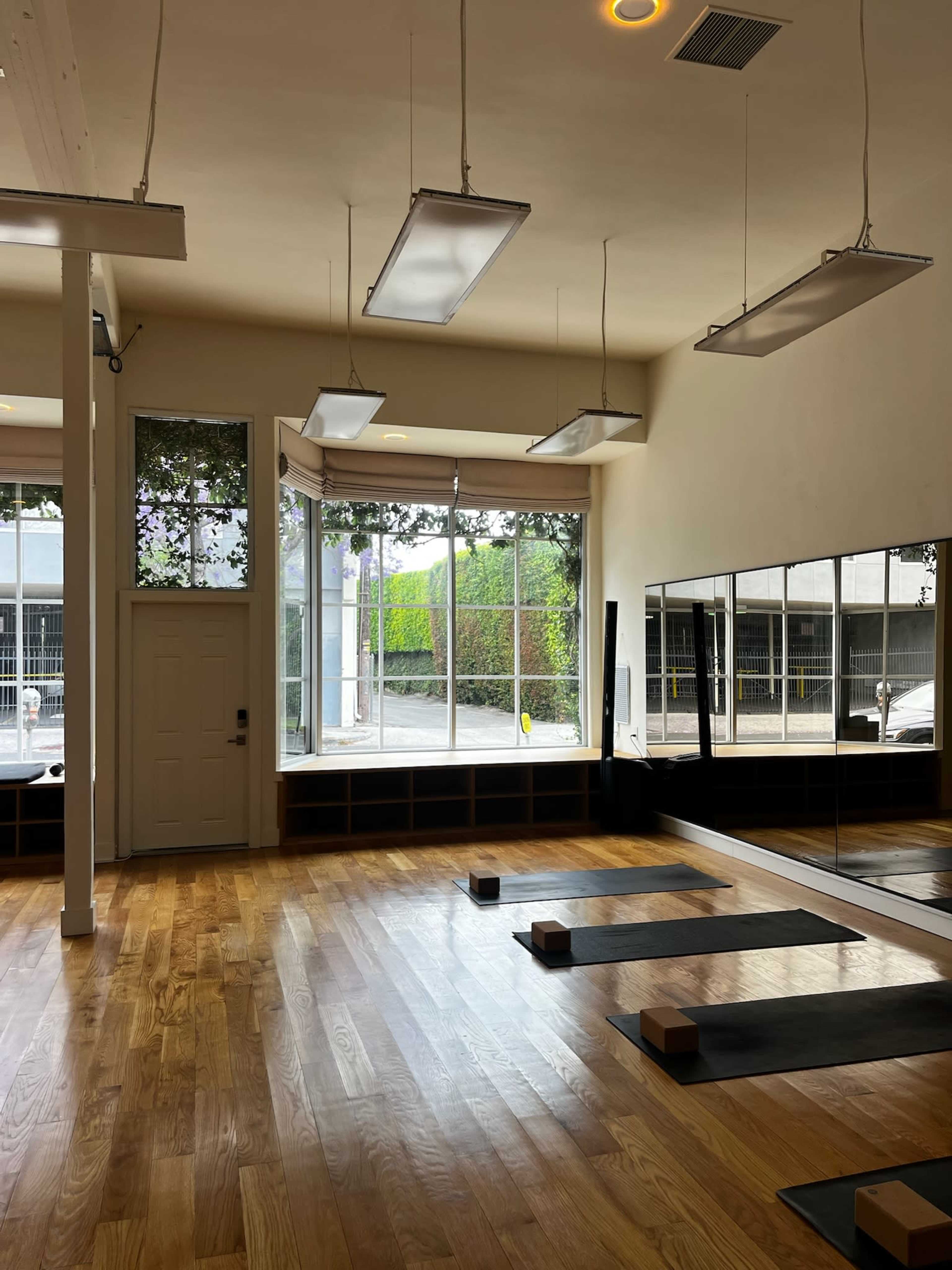 The image shows a spacious yoga studio with wooden floors, large windows, and yoga mats laid out on the floor alongside blocks.