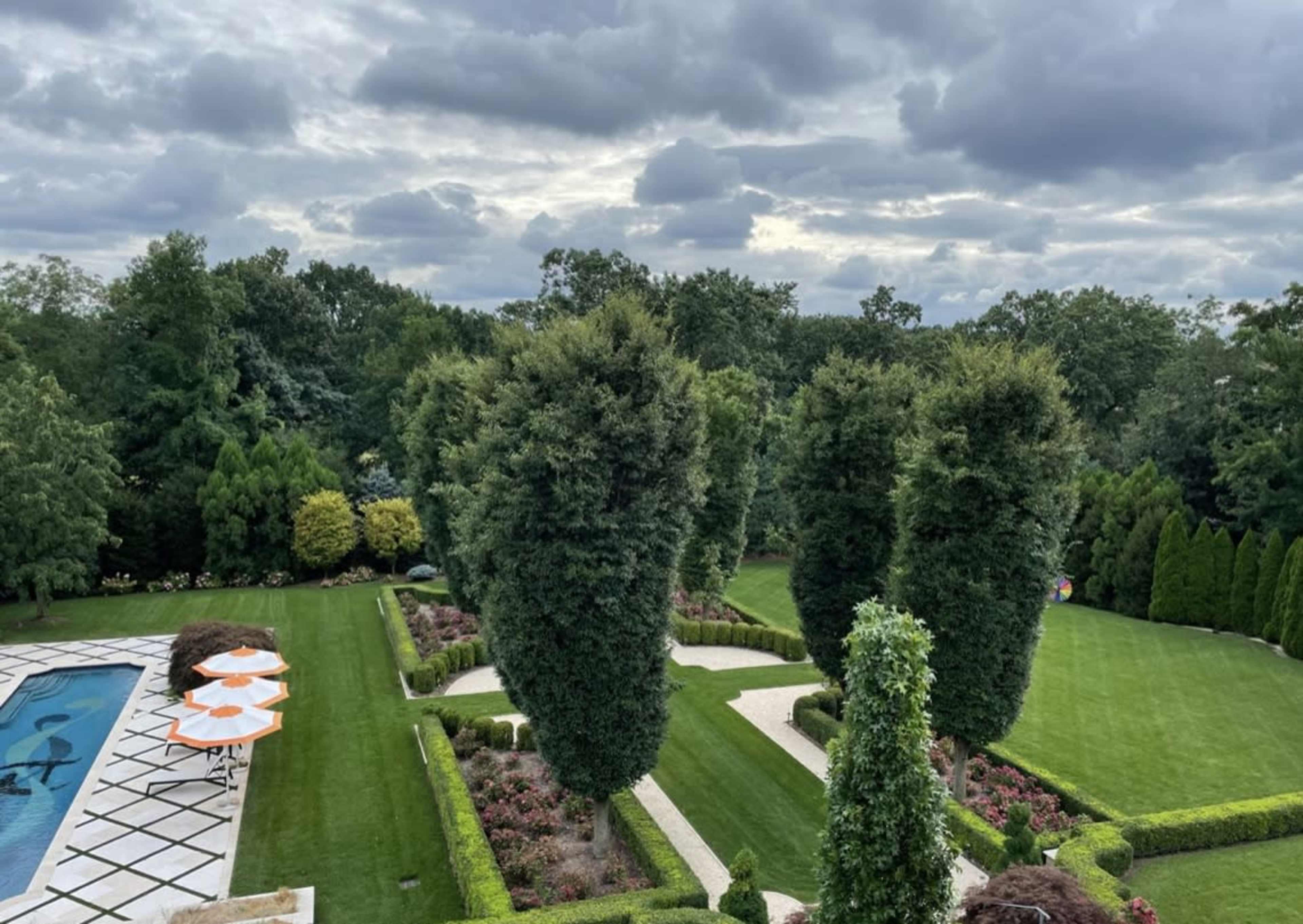 The image shows a well-manicured garden with neatly trimmed hedges, swimming pool, and patio umbrellas under a cloudy sky.
