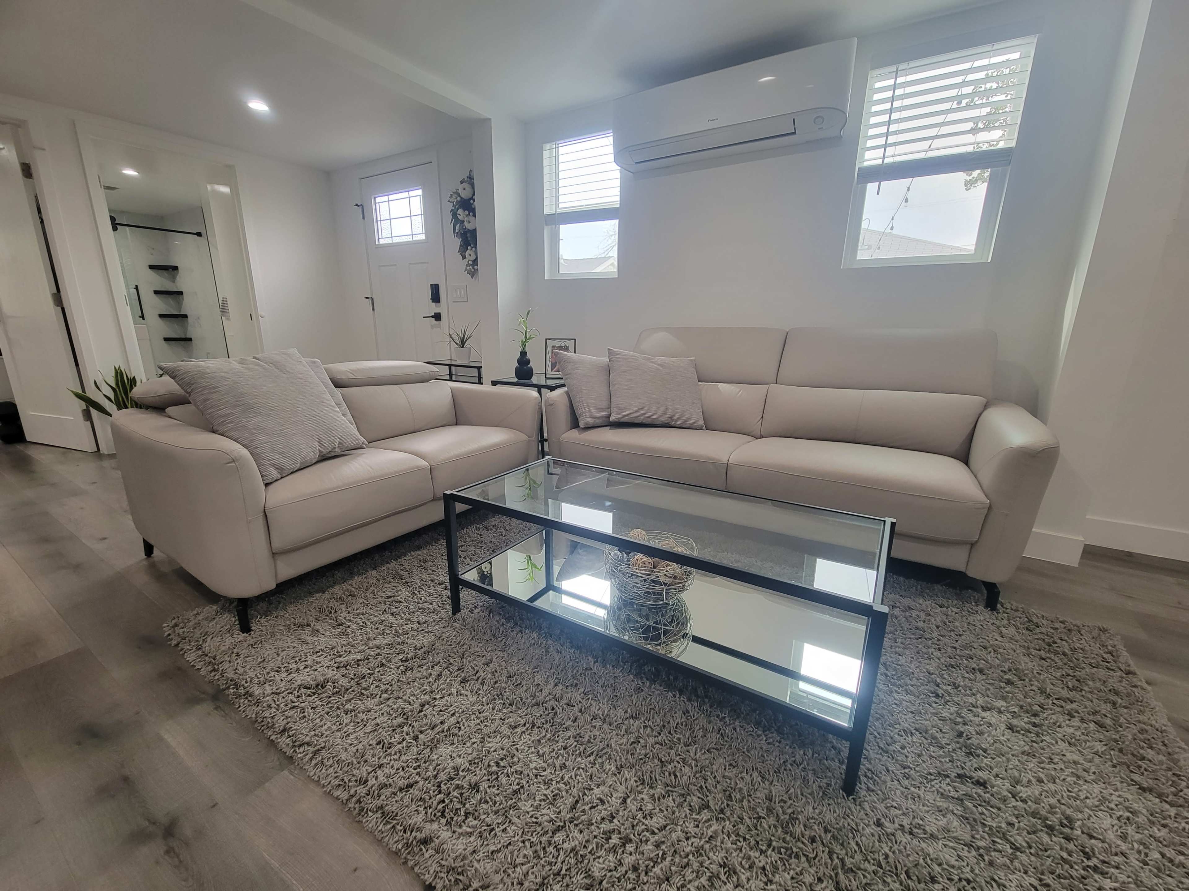 A modern living room with two light-colored sofas, a glass coffee table, and a textured area rug, all situated in a well-lit space.