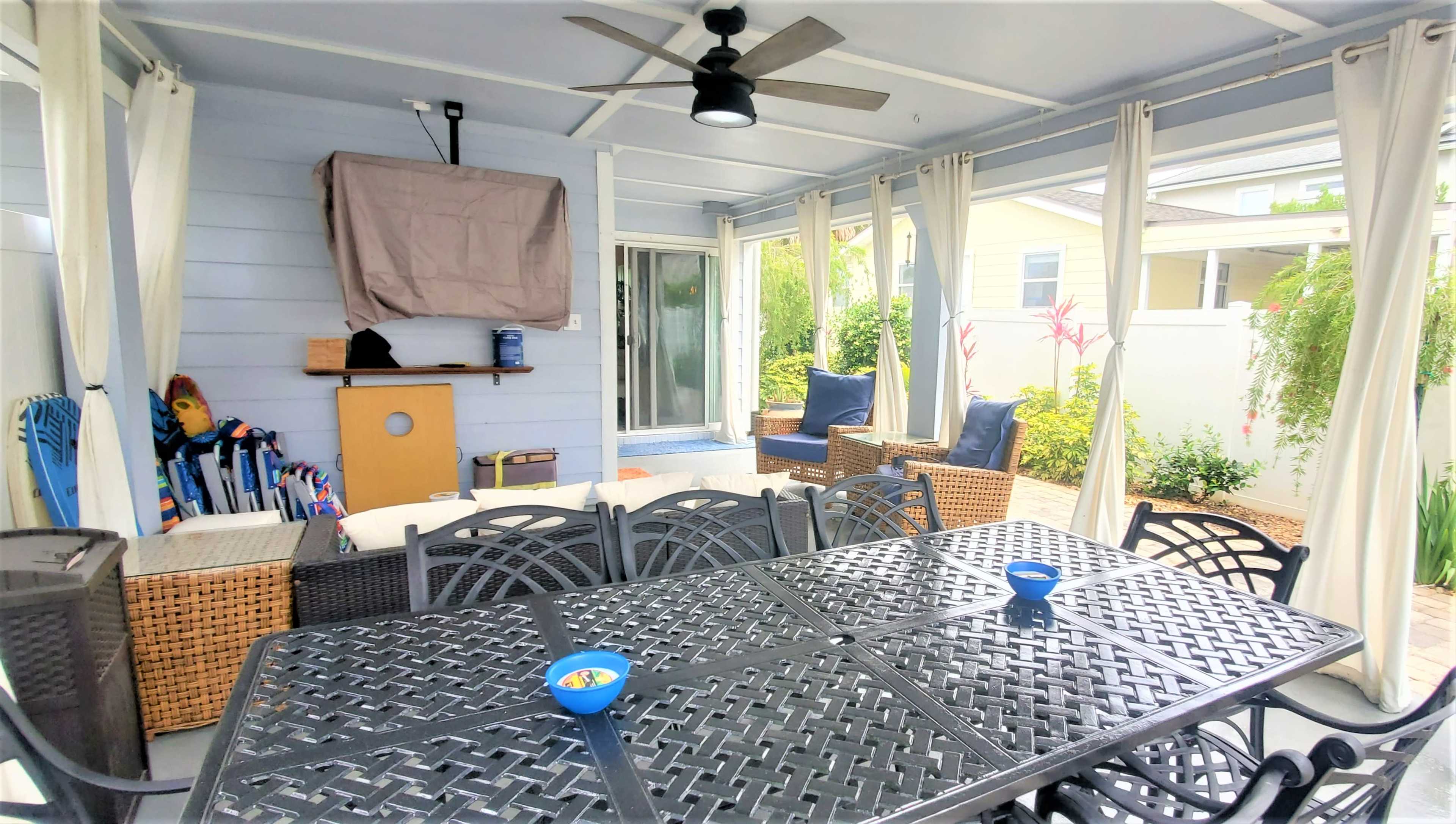 A covered patio area furnished with a large black table, chairs, and a few blue outdoor cushions, surrounded by light-colored curtains and plants.