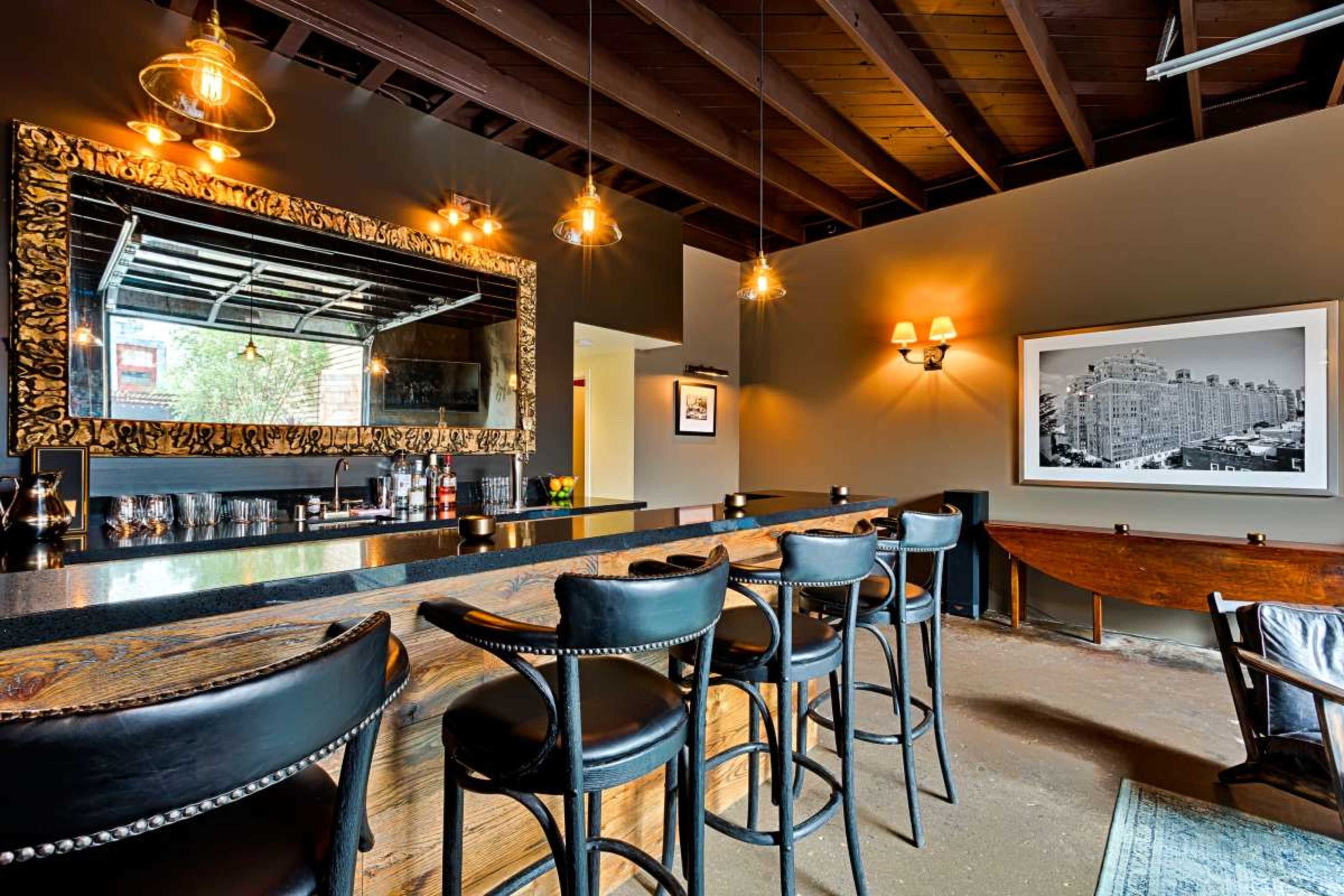 The image shows a modern bar with a black countertop, high-backed chairs, a large decorative mirror, and pendant lighting in a dimly lit room.