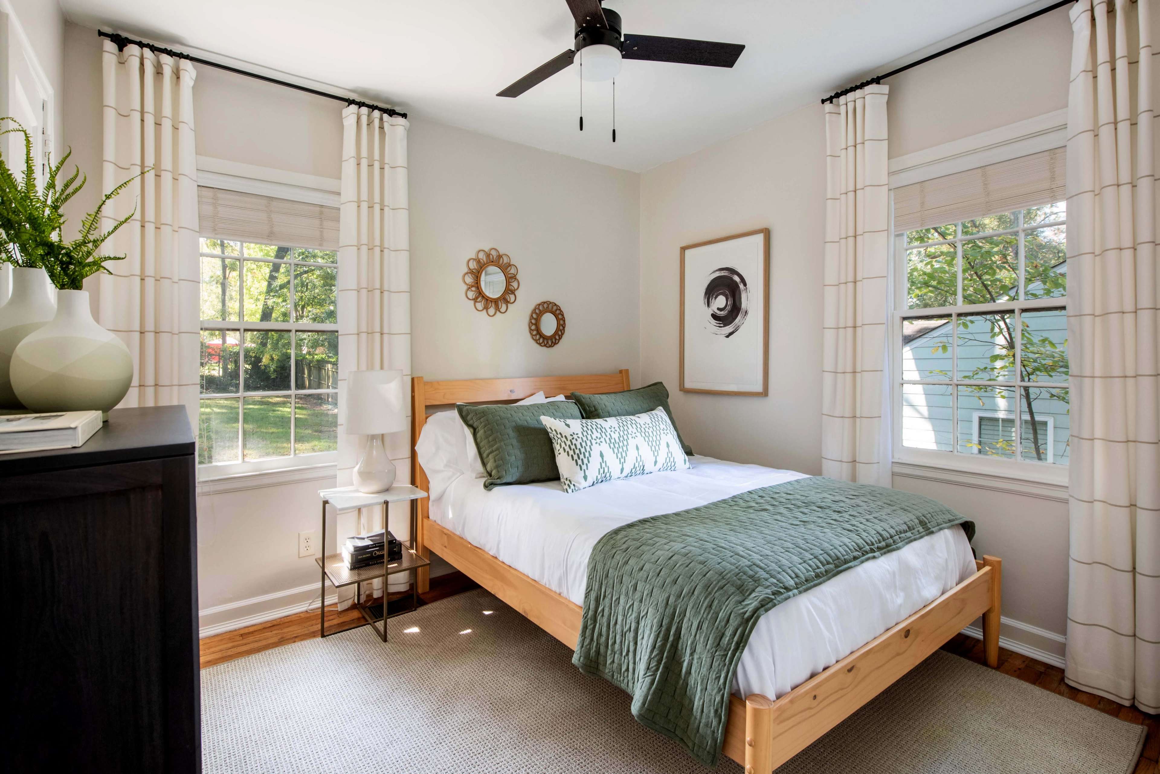 A neatly arranged bedroom featuring a wooden bed with green and white bedding, light-colored walls, and large windows with sheer curtains.