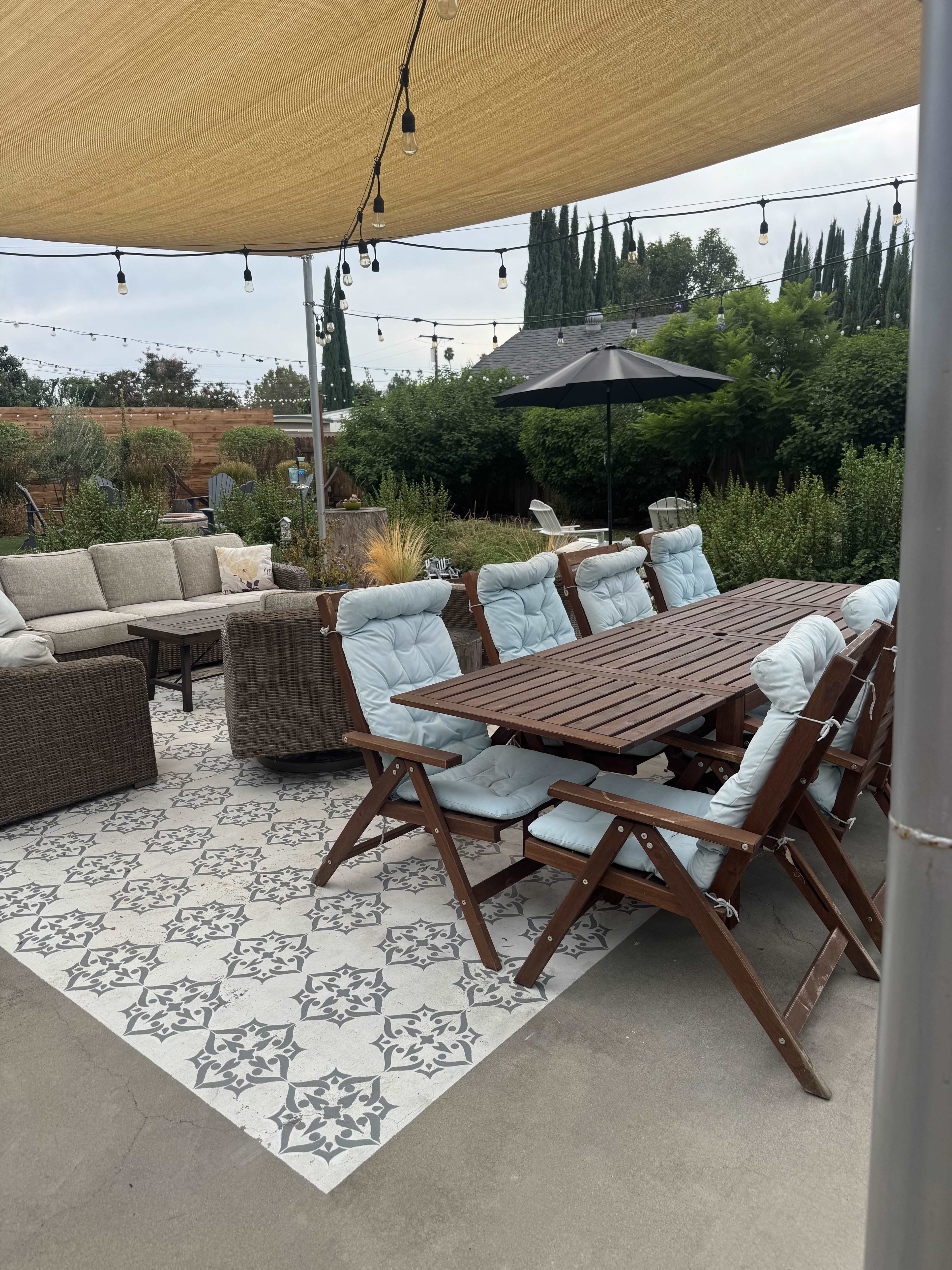 The outdoor patio features a large dining table with wooden chairs and a cozy seating area with upholstered furniture, all shaded by a canopy and surrounded by greenery.