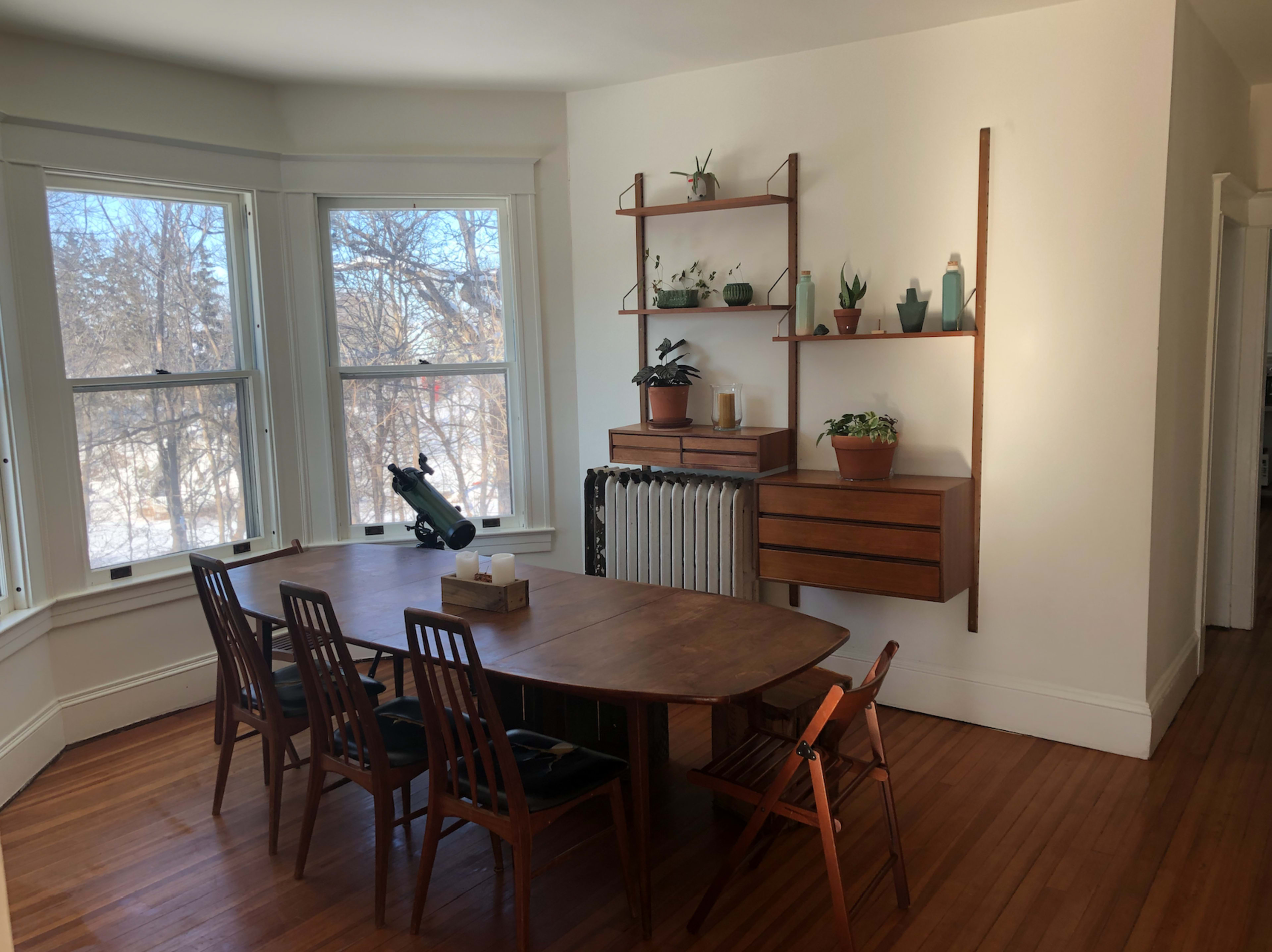 A wooden dining table with six chairs is positioned in a bright room featuring large windows and a wall-mounted shelf with various plants and decorative items.