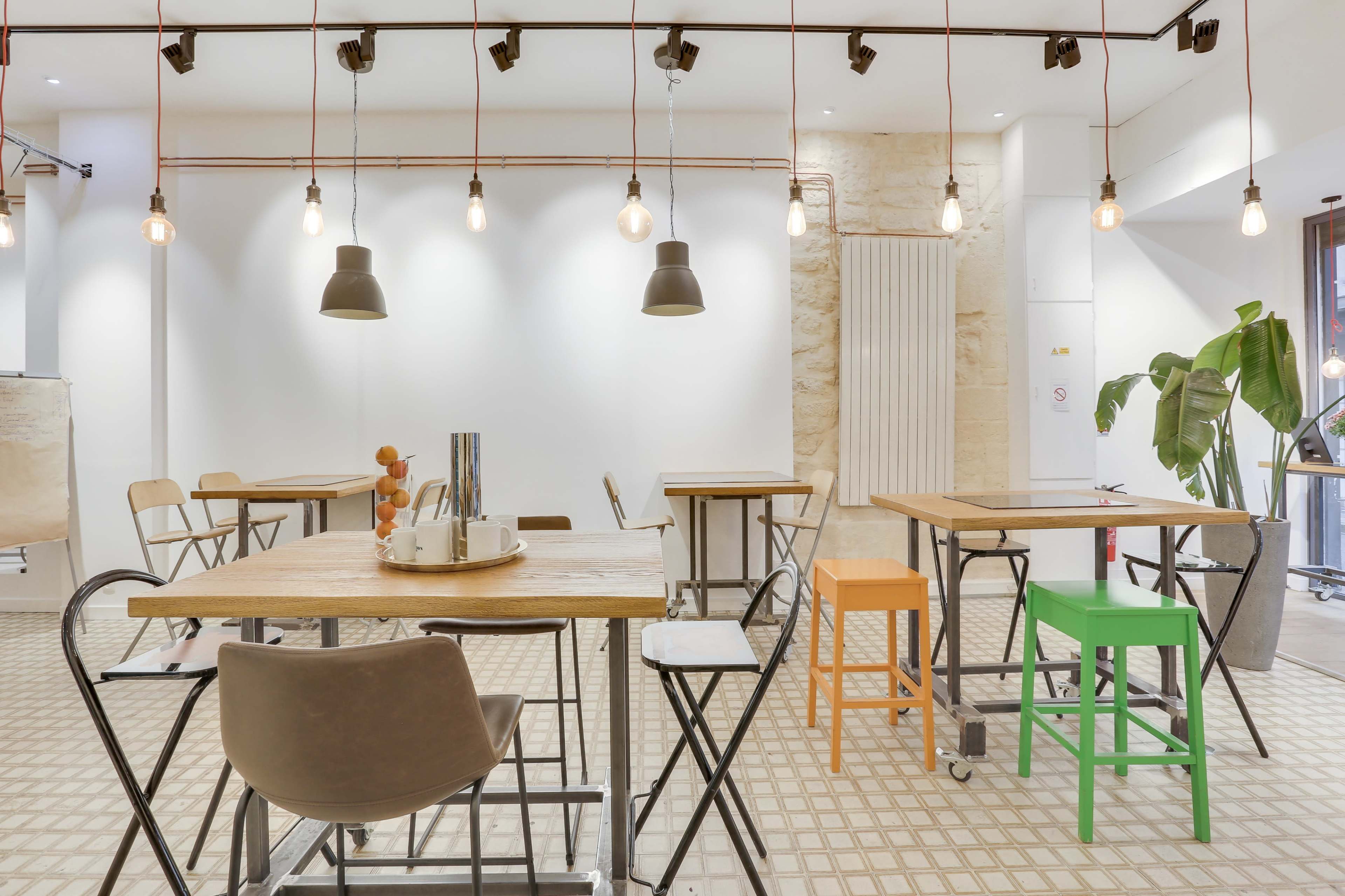 A modern, brightly lit cafe space features wooden tables, various chairs, and pendant lighting hanging from the ceiling.