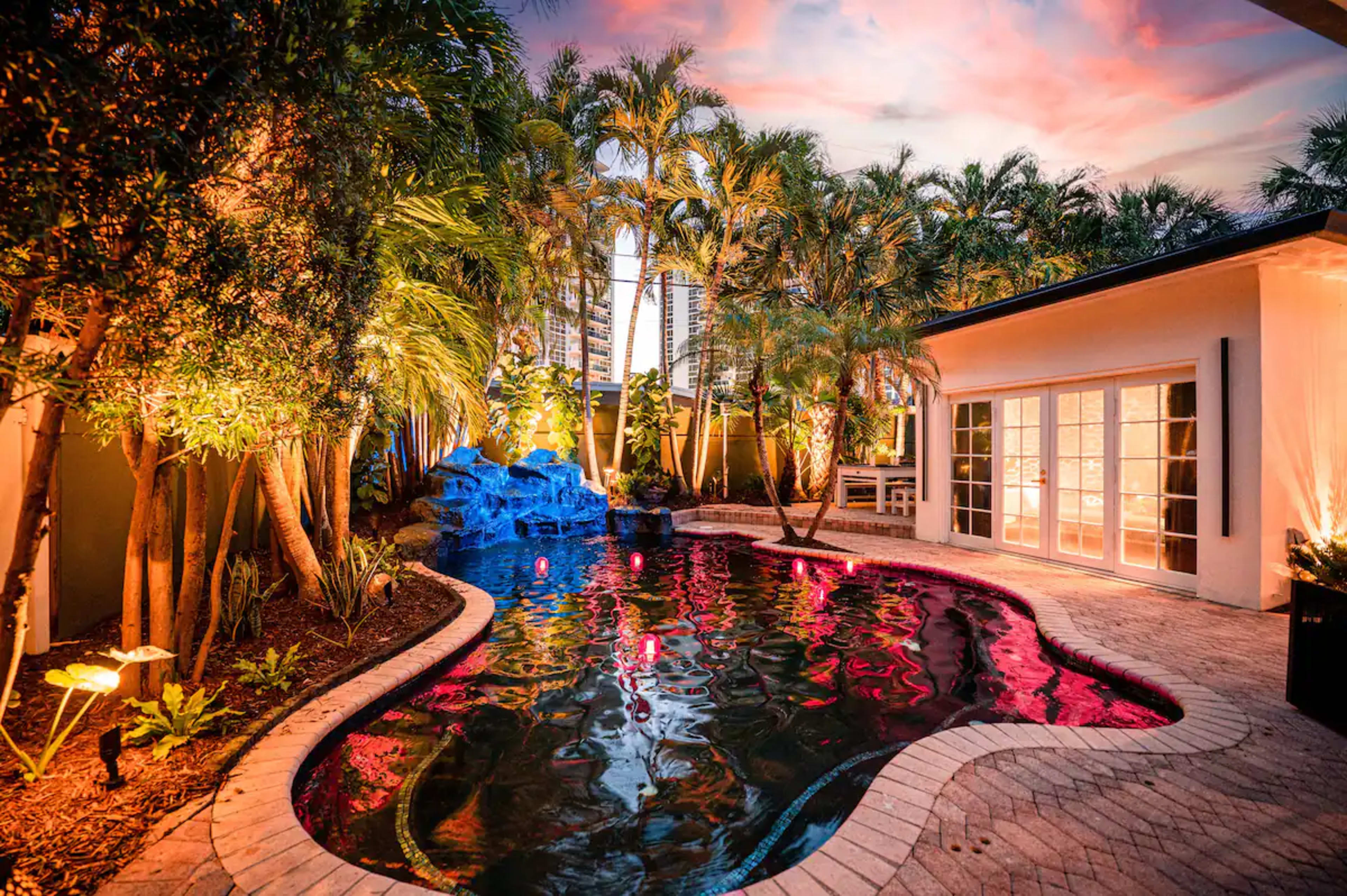 A landscaped backyard with a swimming pool, surrounded by palm trees and illuminated by colorful lights at dusk.