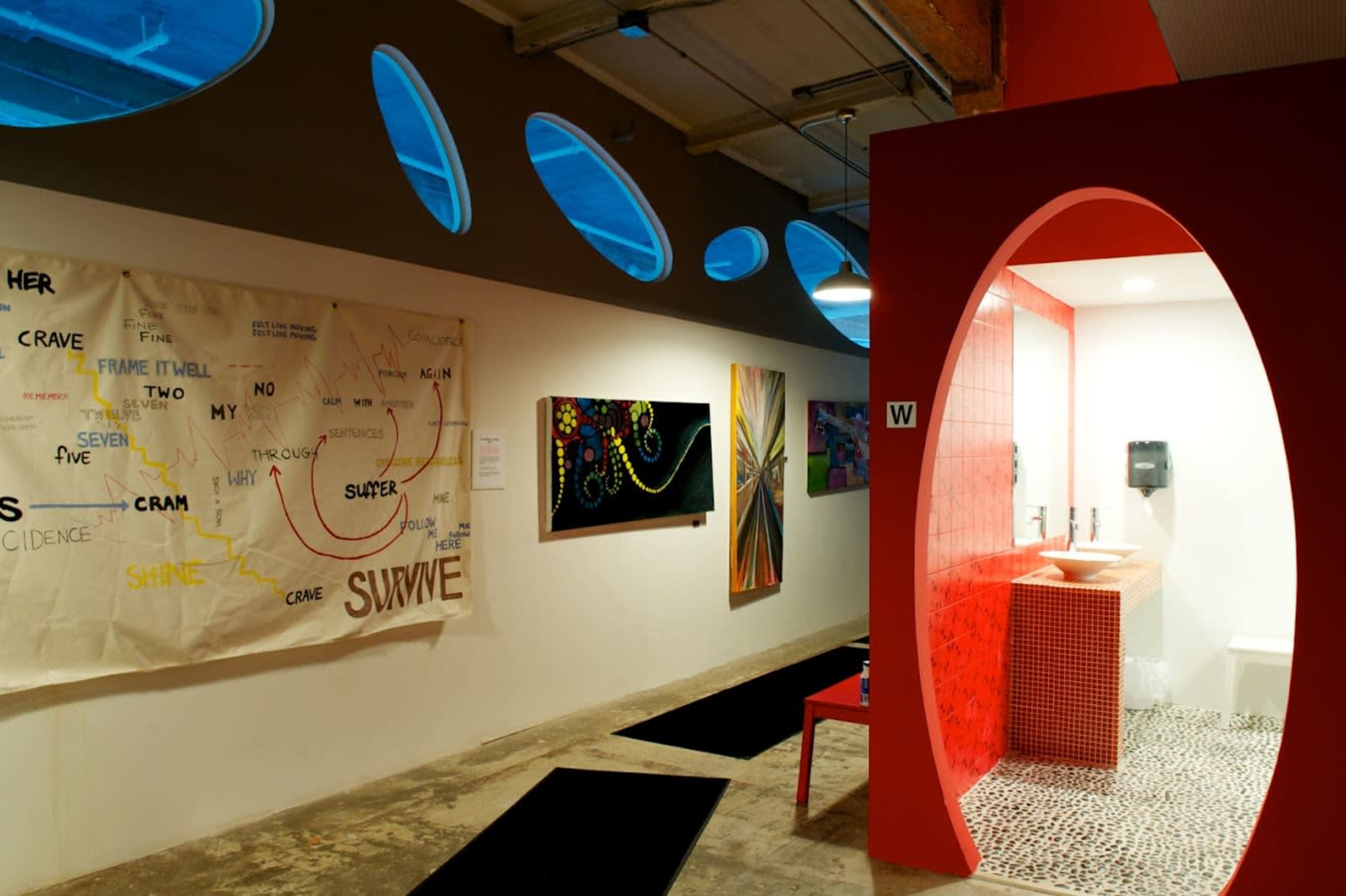 An art gallery with a unique restroom design, featuring a red wall structure and various artworks displayed along the walls.