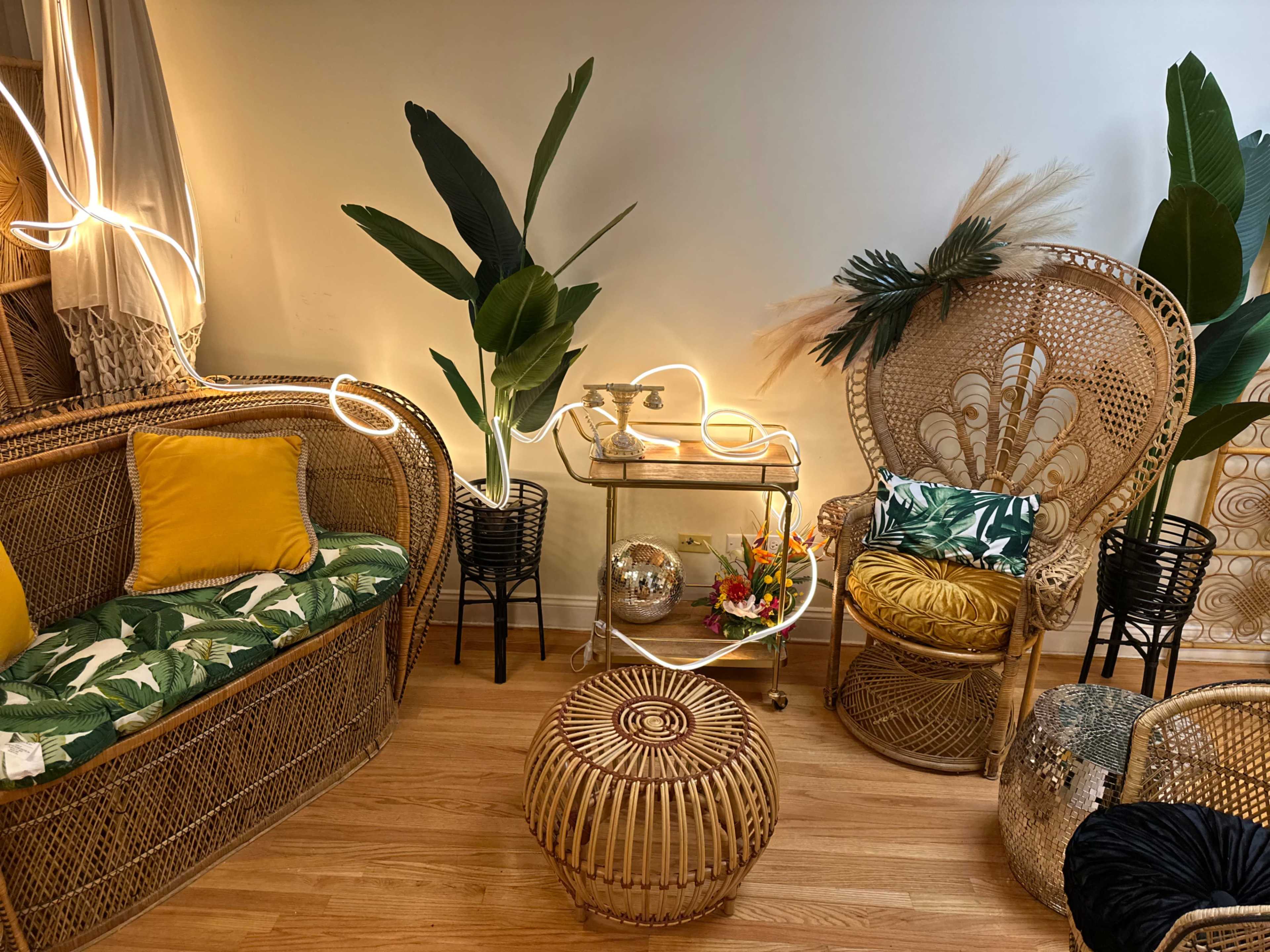 The image features a cozy interior space with rattan furniture, including a sofa adorned with yellow and patterned cushions, a decorative side table, and a round woven stool, complemented by tropical plants and soft lighting.