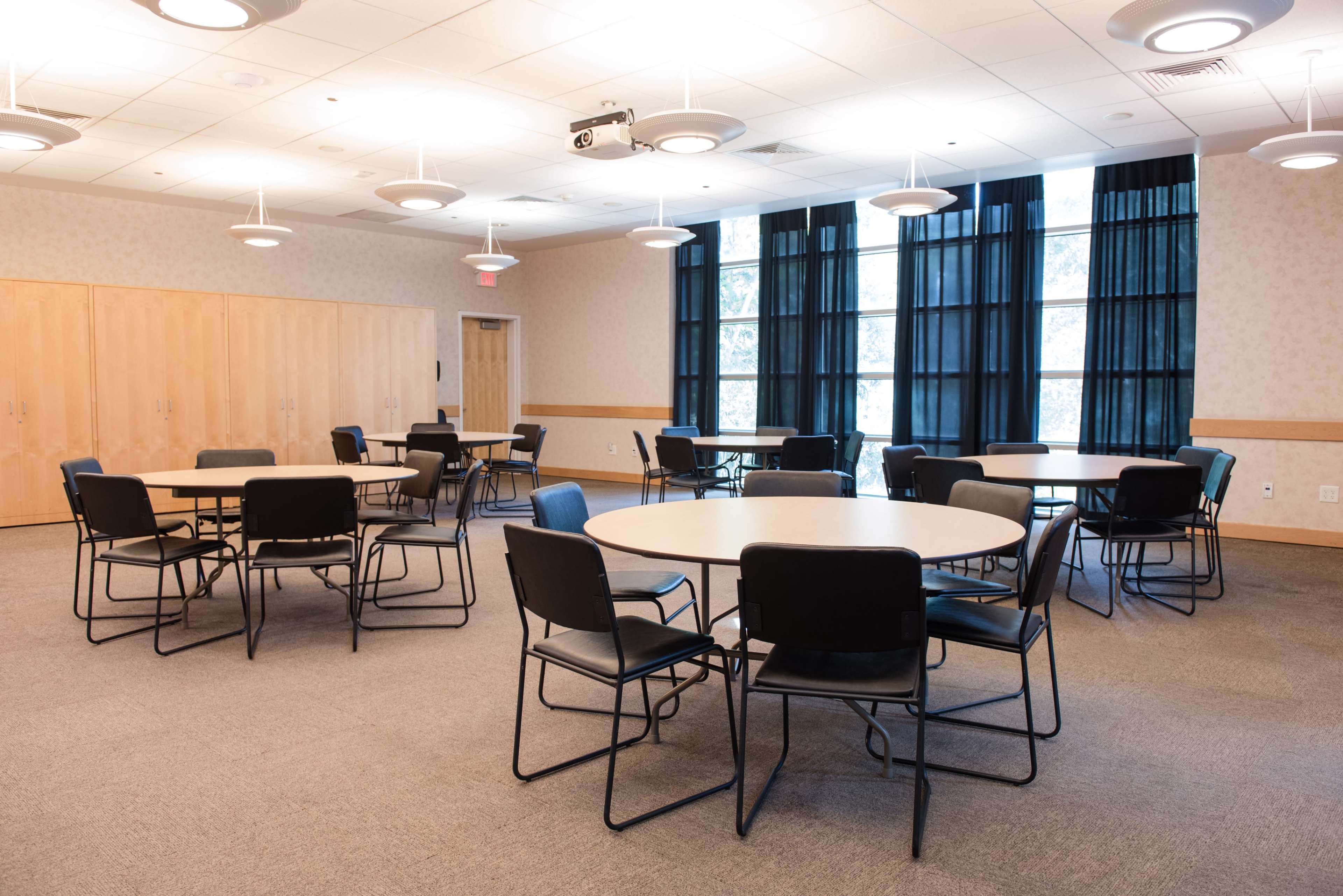 A large meeting room features several round tables surrounded by chairs, with windows covered by black curtains.