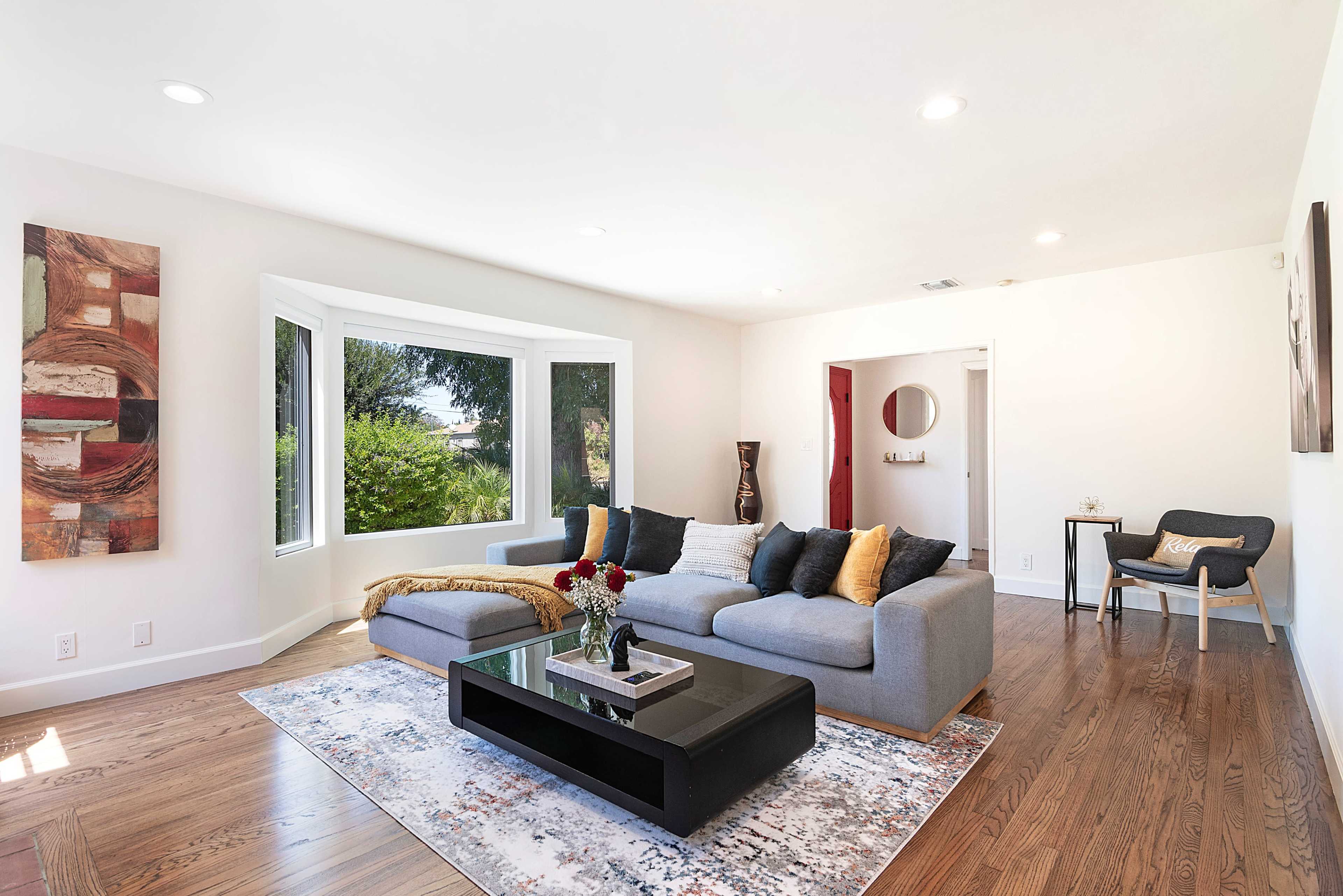 A spacious living room features a gray sectional sofa, a black coffee table, and large windows with views of greenery.