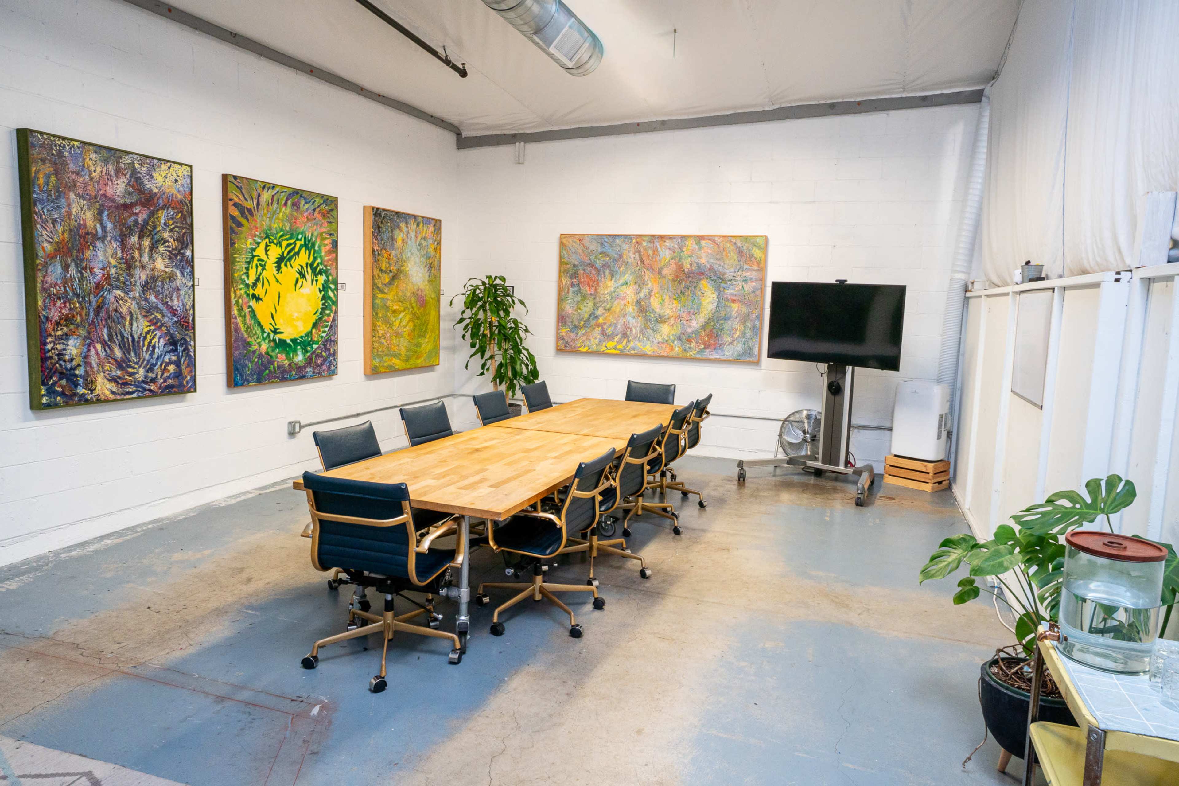 The image depicts a modern conference room featuring a long wooden table surrounded by black office chairs, colorful abstract paintings on the walls, a large television screen, and potted plants.