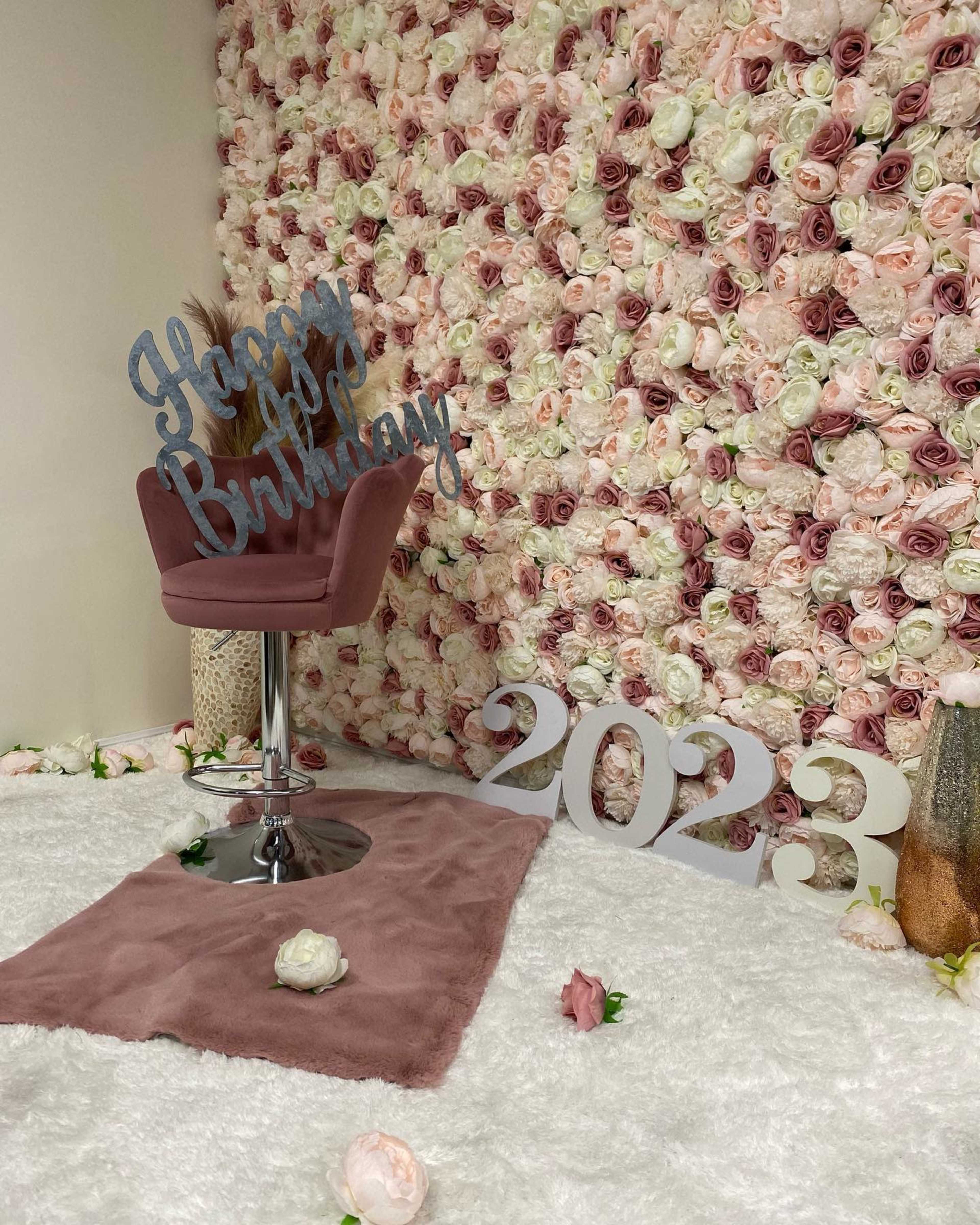 The scene features a chair in front of a wall covered with artificial flowers, accompanied by a decorative sign that reads "Happy Birthday" and the numbers "2023" displayed on the floor.