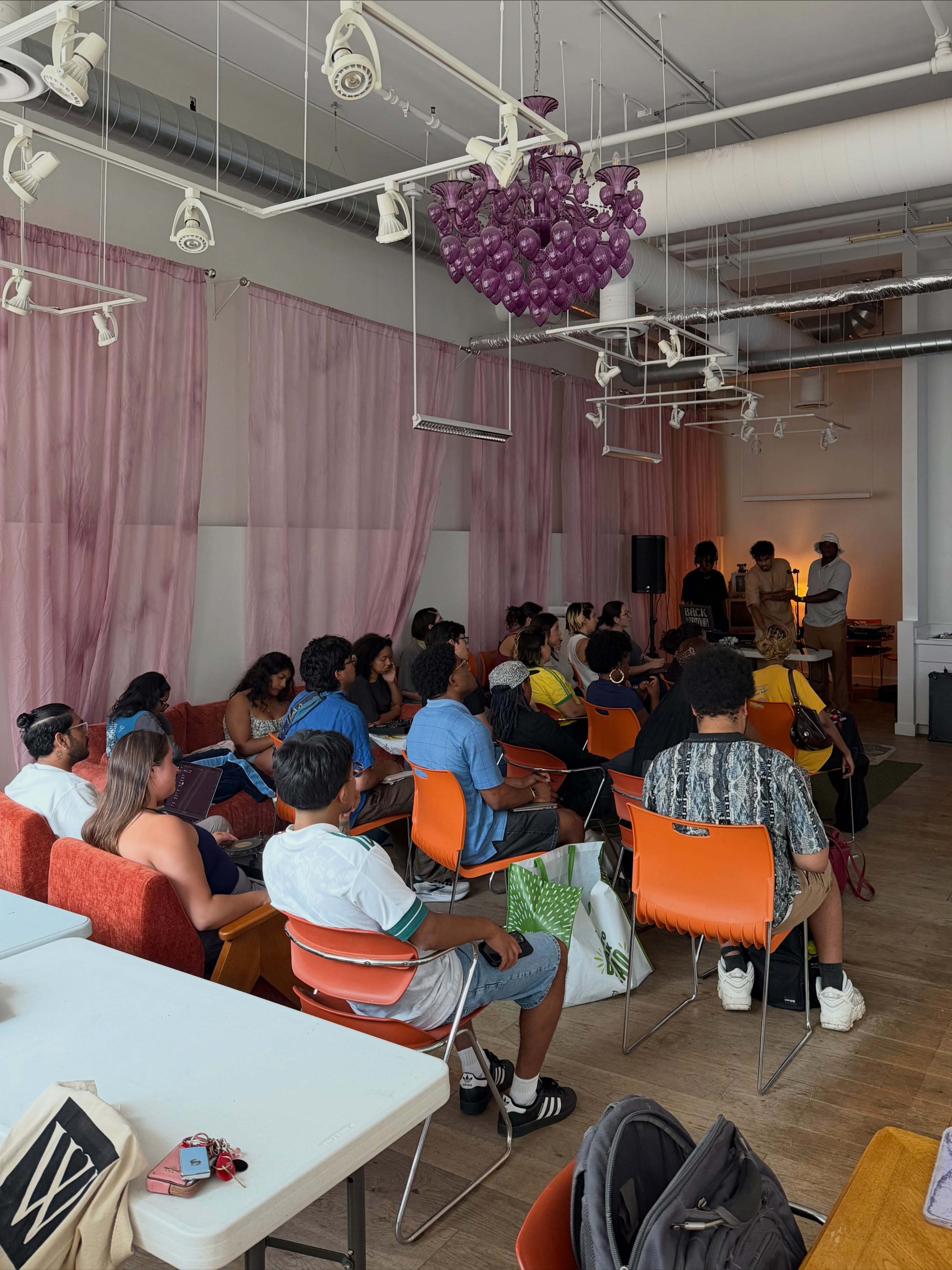 A group of people sits in a brightly lit room with pink curtains and modern furnishings, listening to a presentation.
