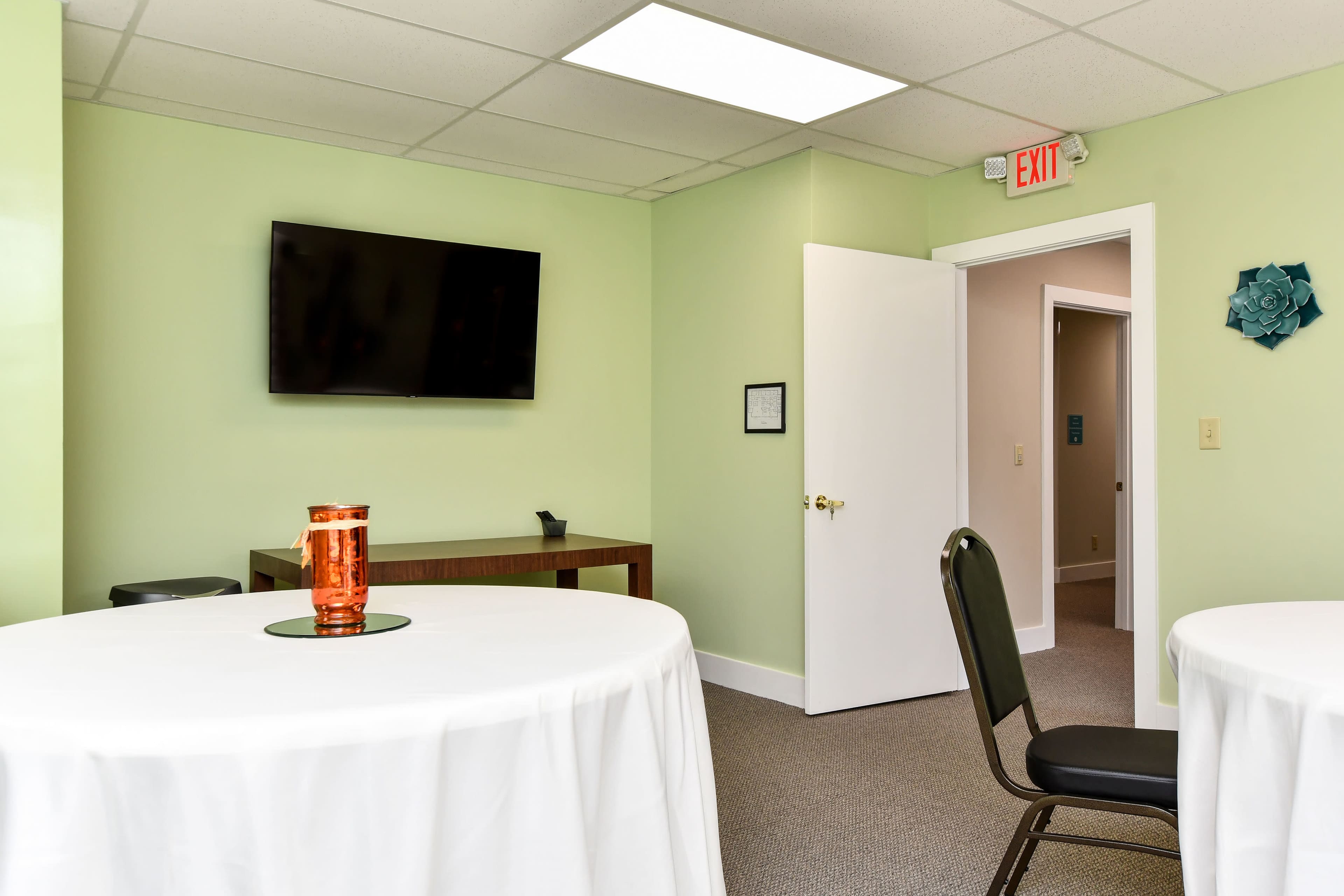 A meeting room with light green walls features two tables covered with white tablecloths, a television mounted on the wall, and a door leading to another area.