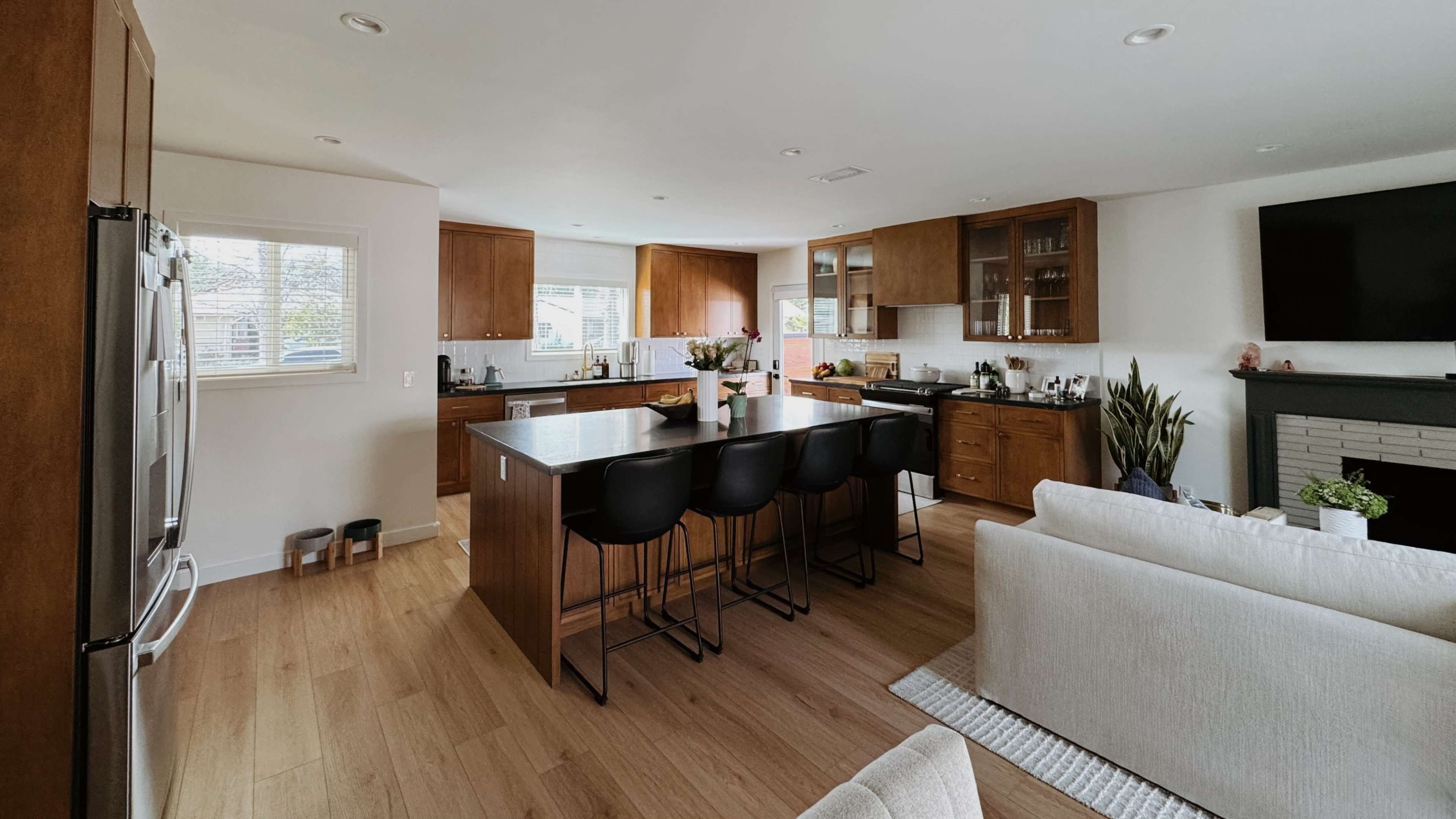 The image shows a modern kitchen with wooden cabinetry, an island with seating, and a connected living area featuring a light-colored sofa.