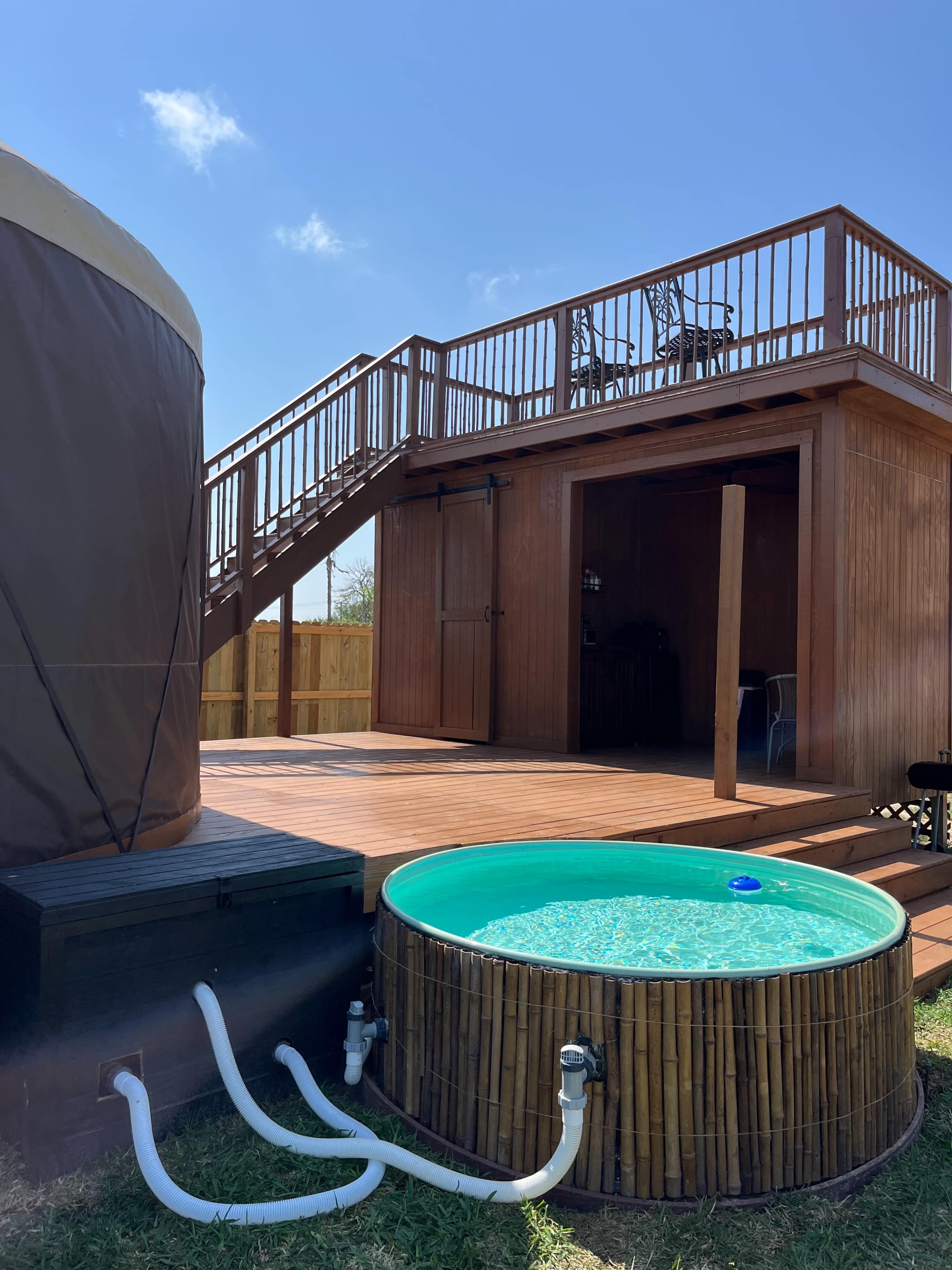 A wooden deck features a round, above-ground pool next to a yurt and a staircase leading to a second-floor area.