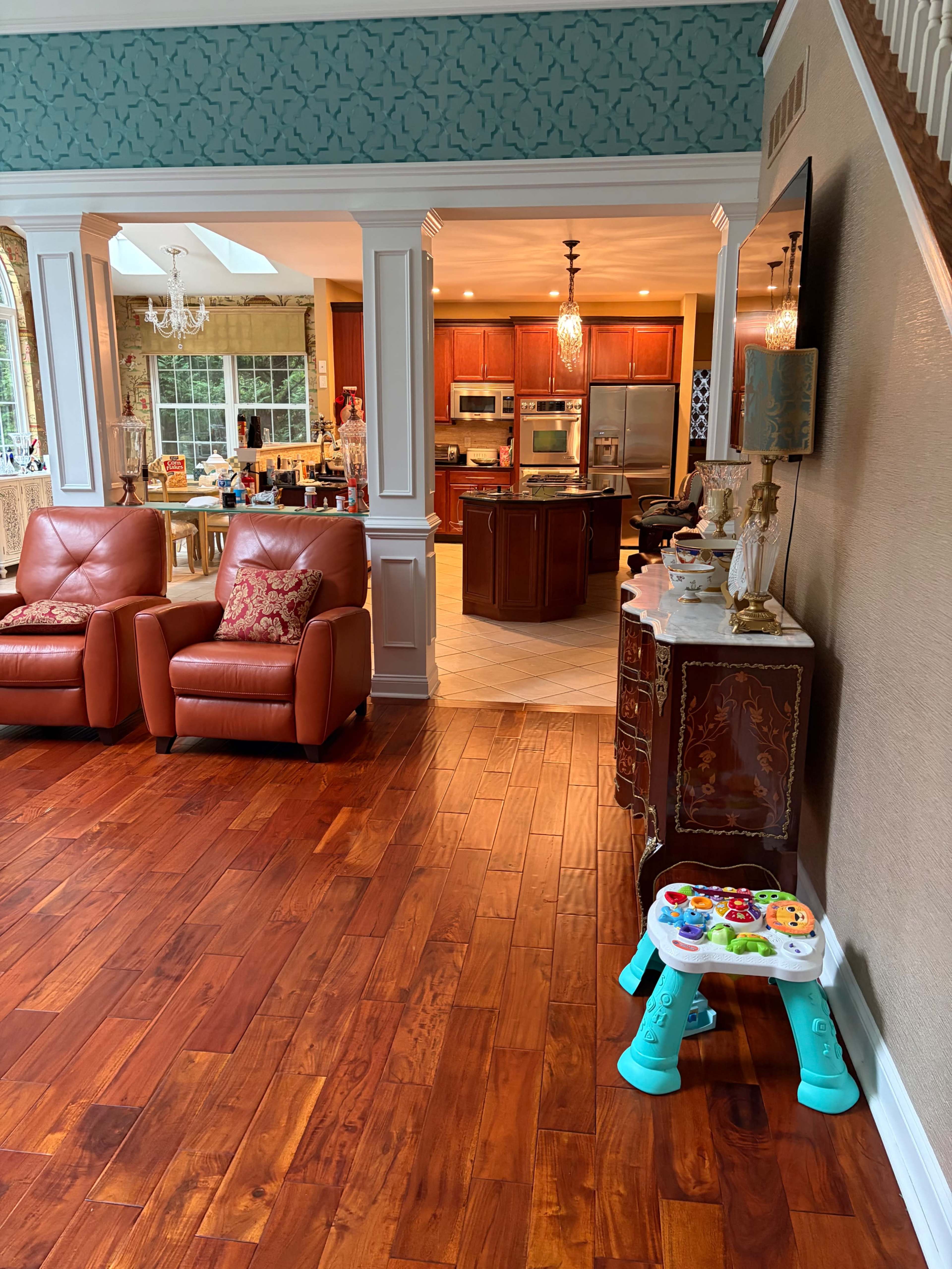 The image shows a spacious living area with wooden flooring, two reddish-brown armchairs, and a view into a kitchen featuring wooden cabinetry.