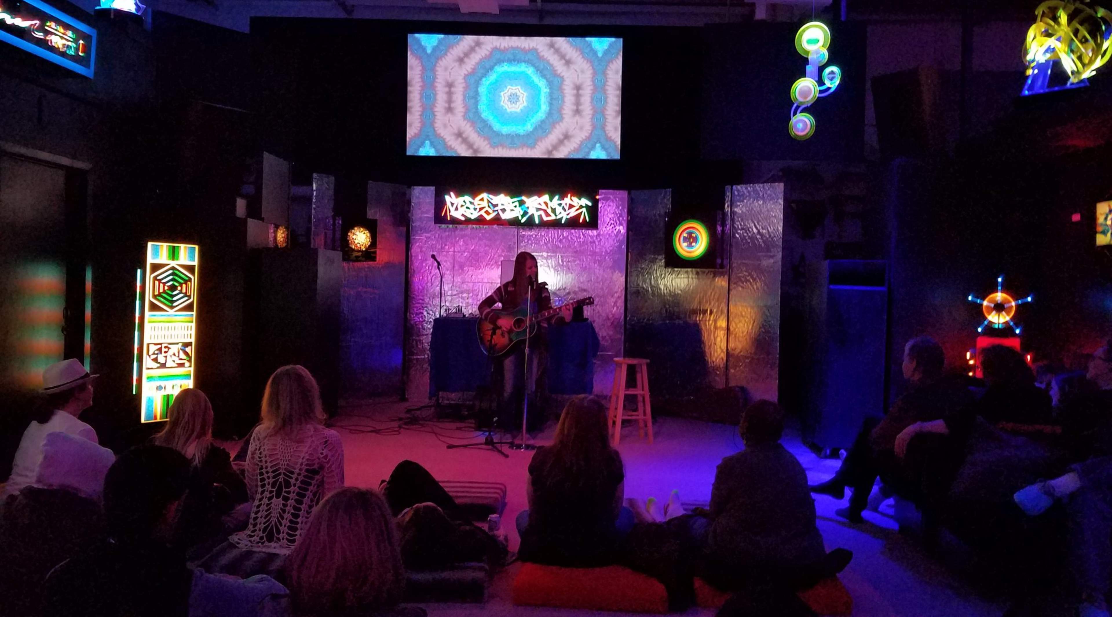 A musician plays guitar on stage in front of an audience, with colorful lights and a patterned projection screen in the background.