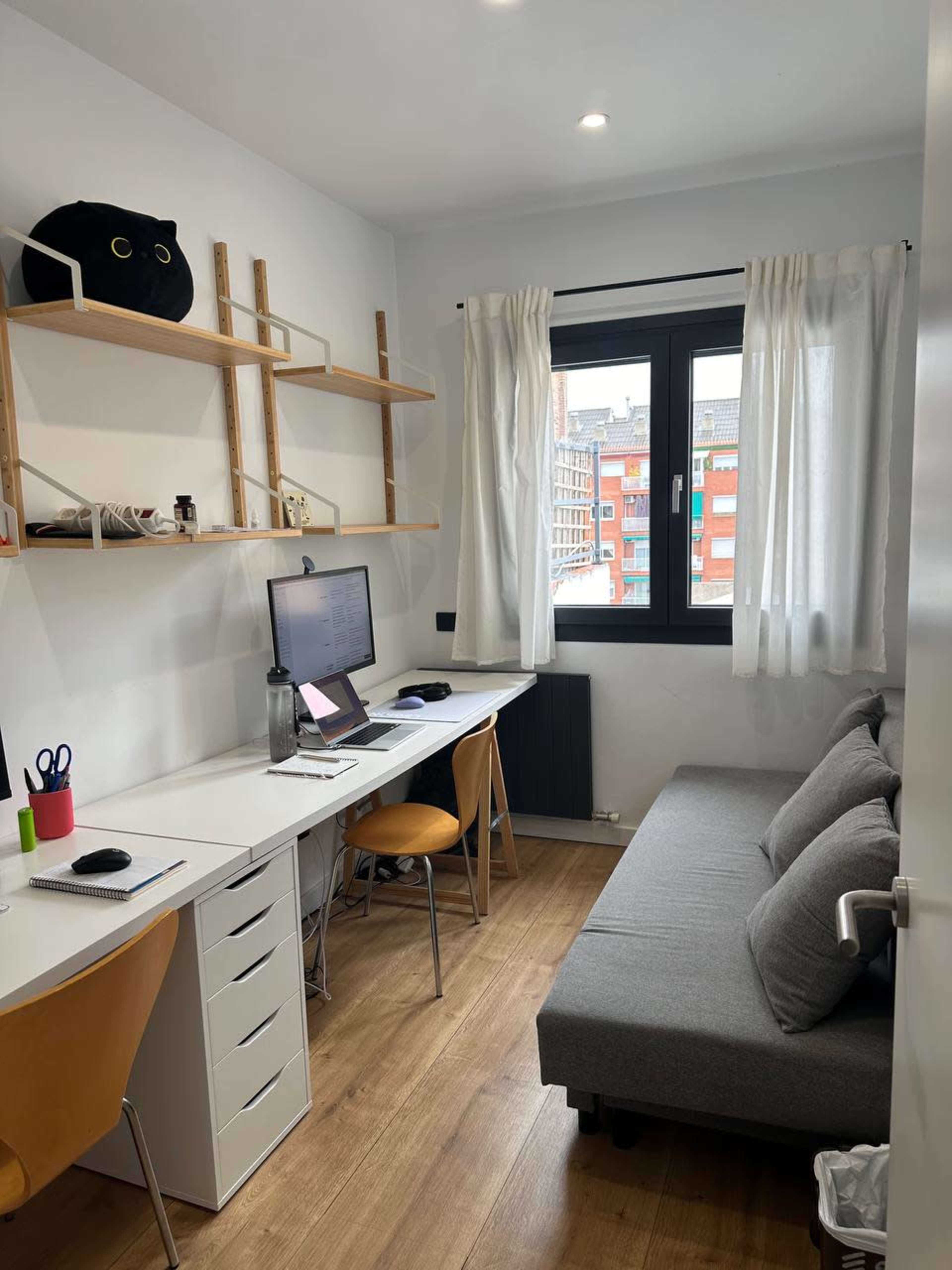 A small home office with a desk, computer, and a gray sofa, featuring white curtains and wooden shelves against the walls.