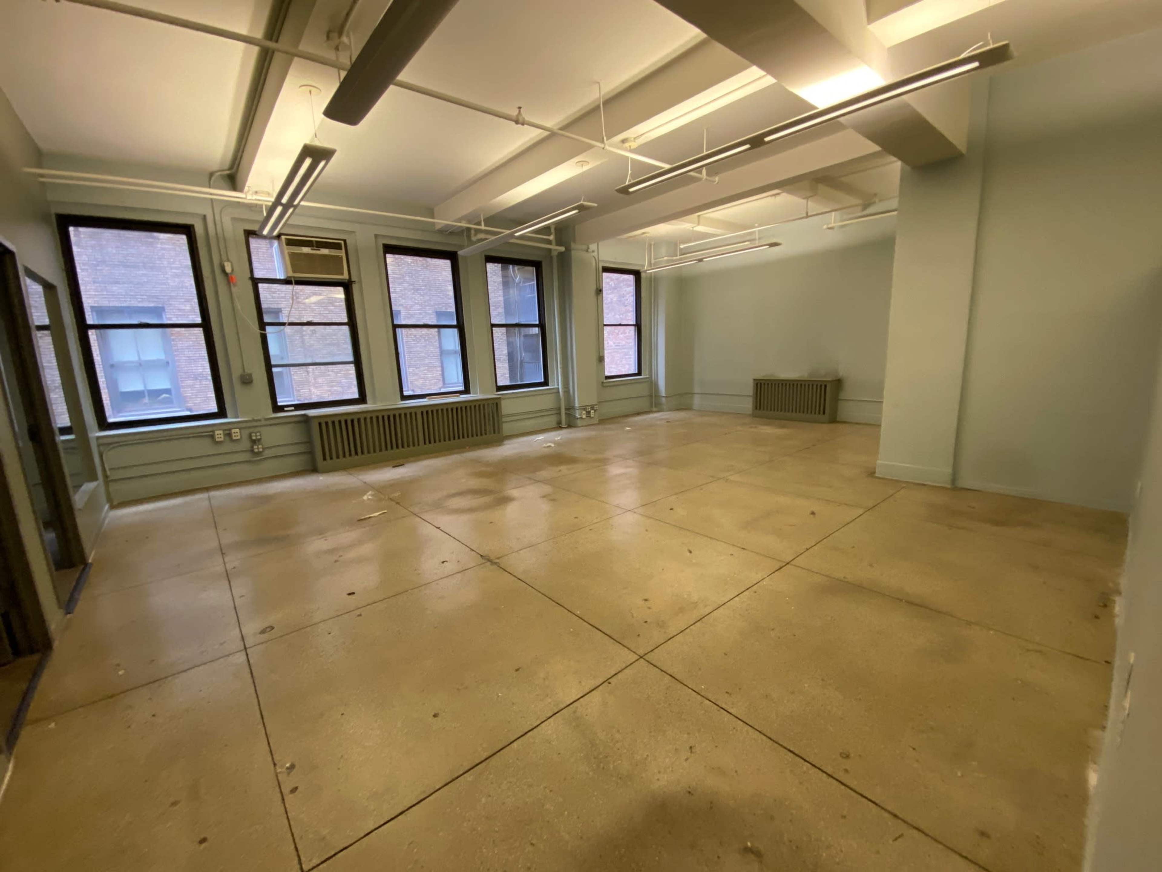 The image shows a vacant room with concrete flooring, large windows, and minimal furnishings.
