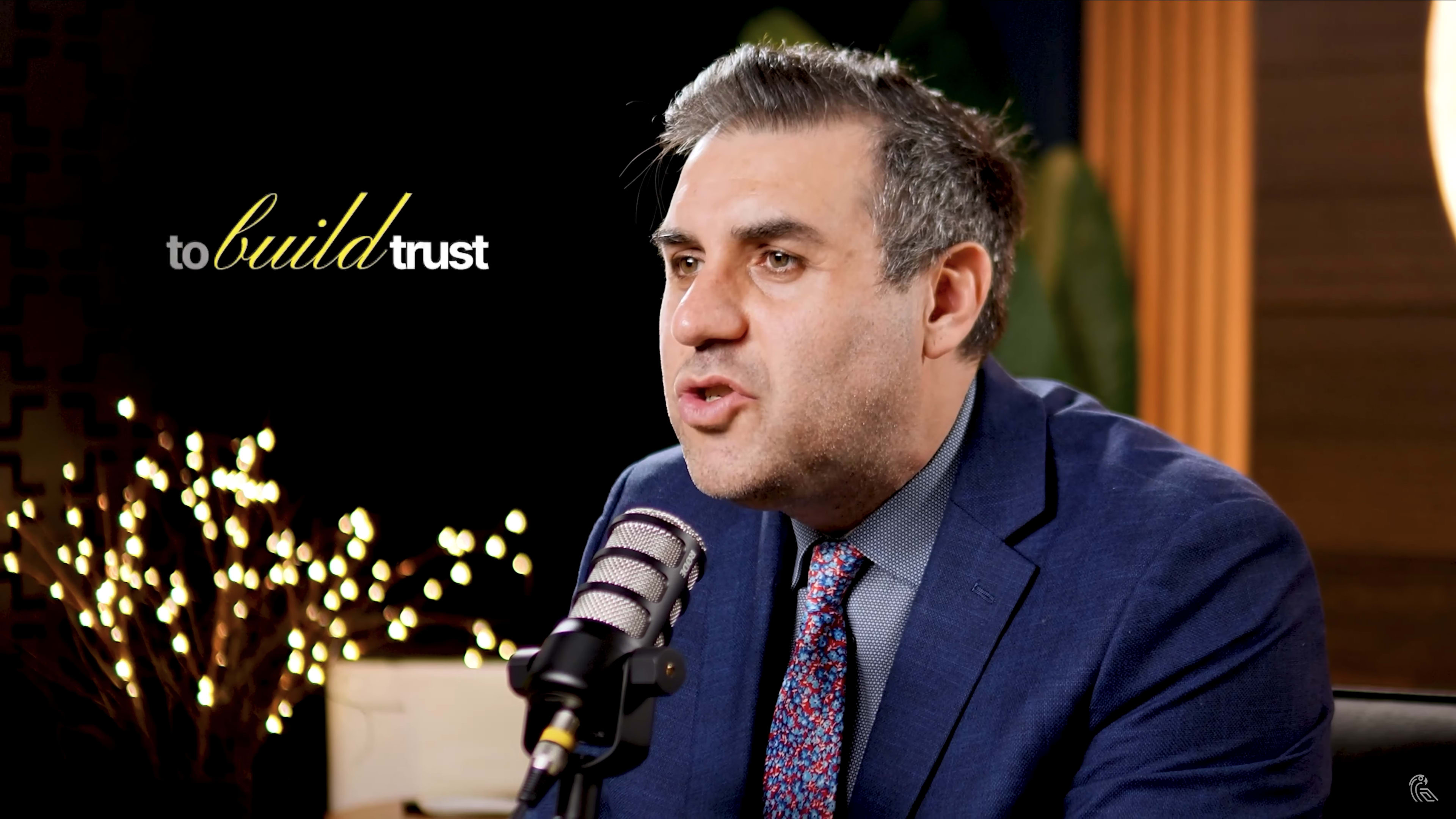A man in a suit is speaking into a microphone in a studio setting with soft lighting and decorative plants in the background.