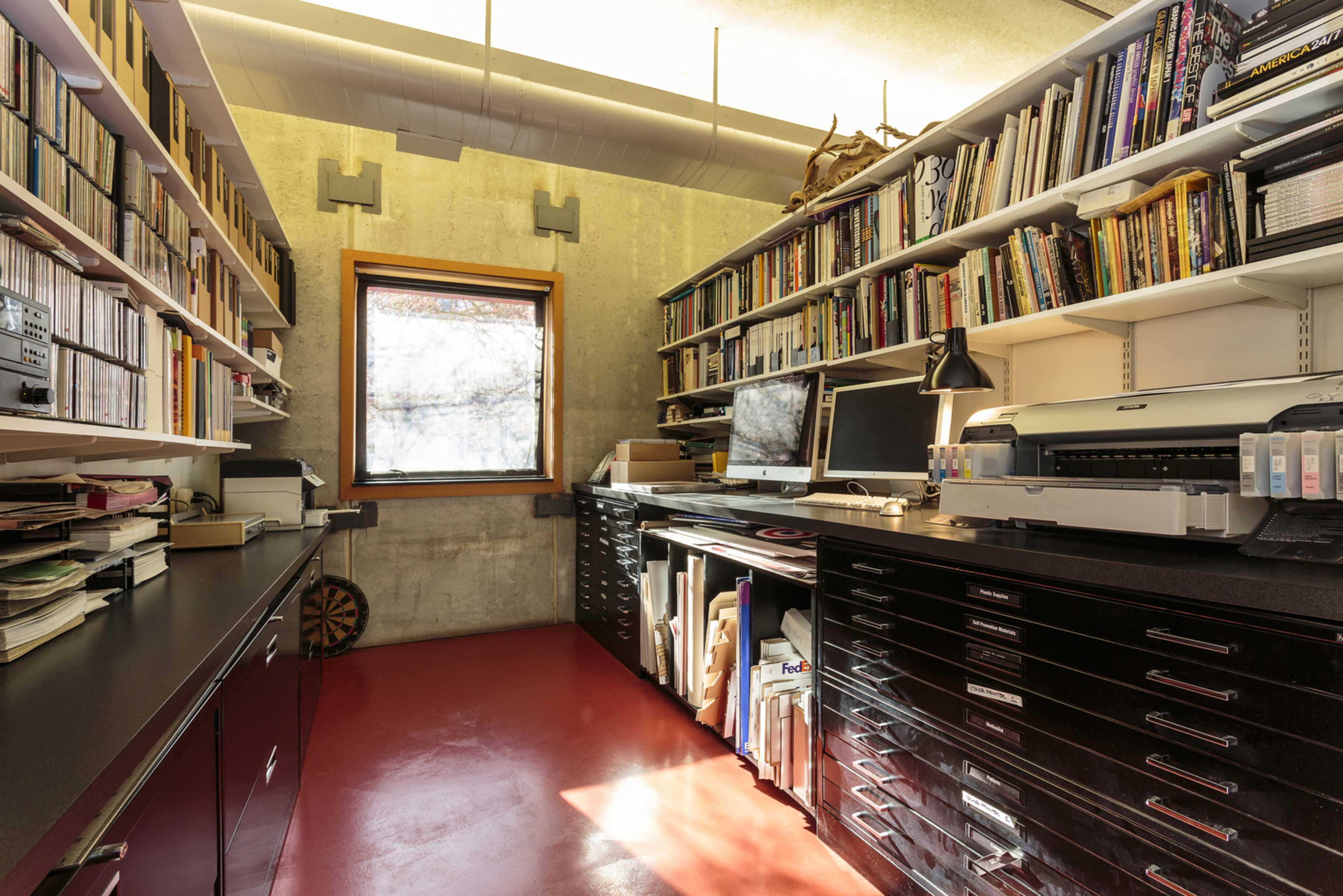 A compact office space features shelves filled with books and CDs, a desk with computers and a printer, and a window providing natural light.