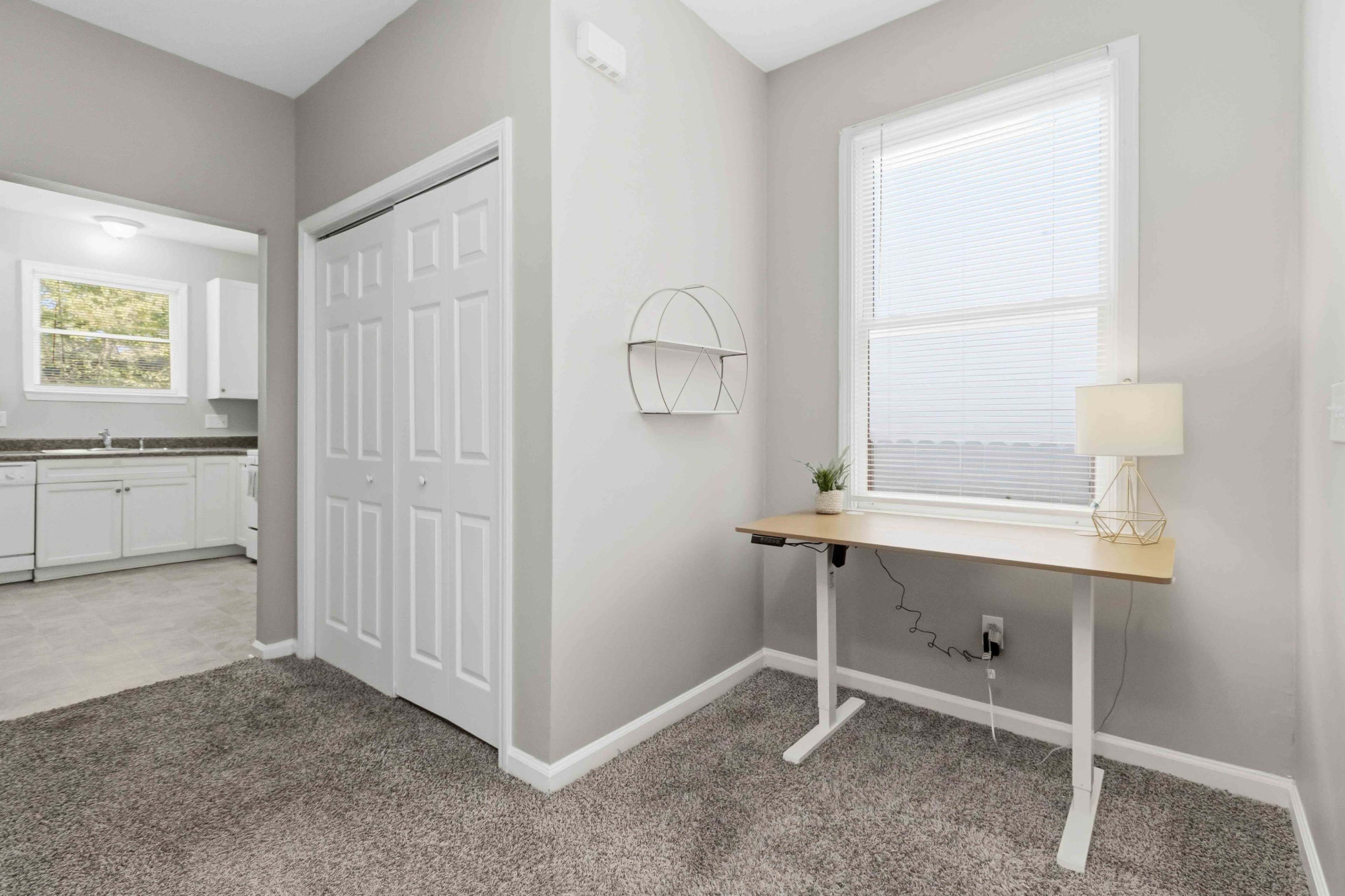 The image shows a light-colored room featuring a small desk with a lamp, a closet with double doors, and a window providing natural light.