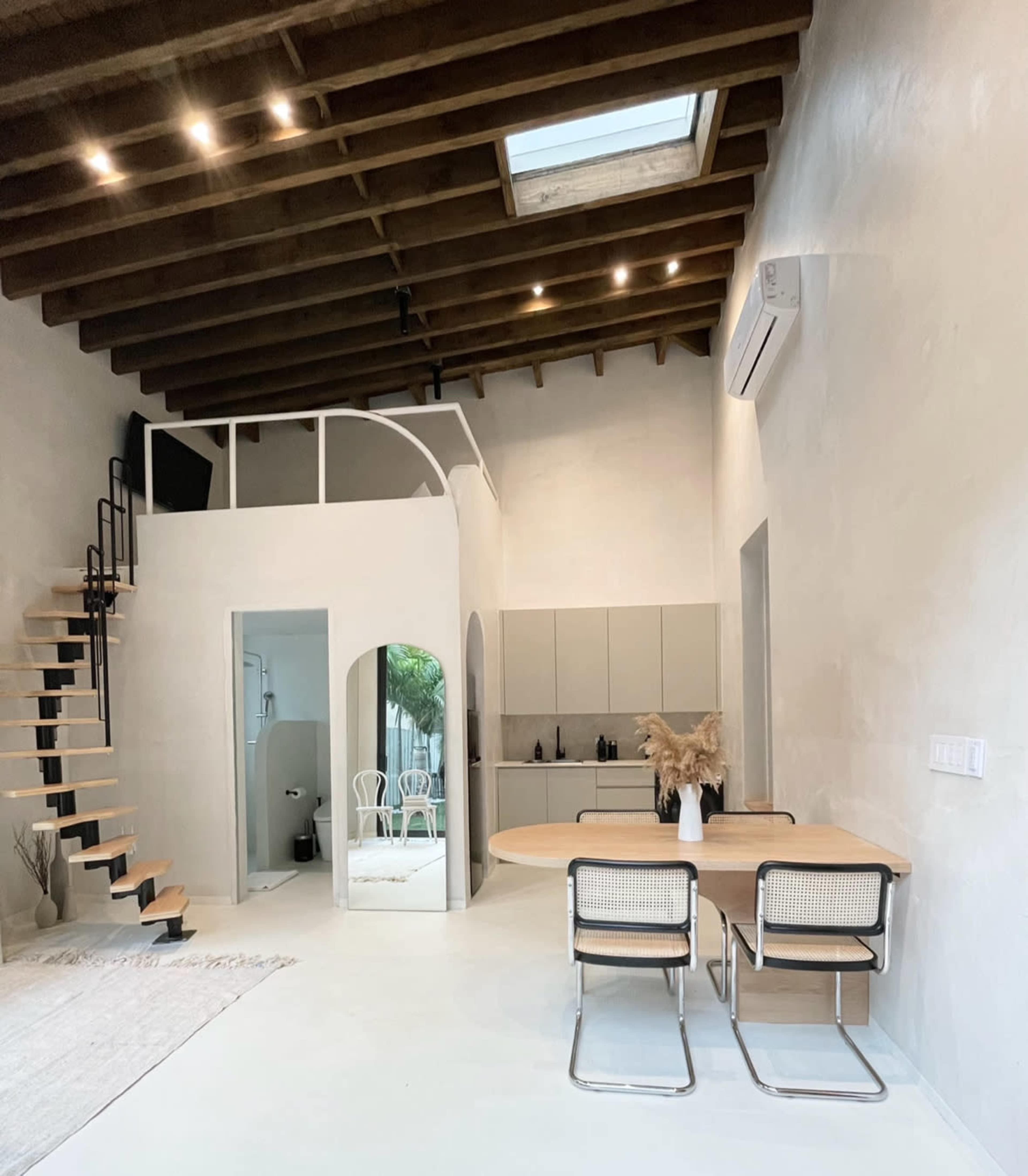 The image shows a modern interior space featuring a staircase, a dining area with a round table and chairs, and a kitchen area, all under a high ceiling with exposed wooden beams and a skylight.
