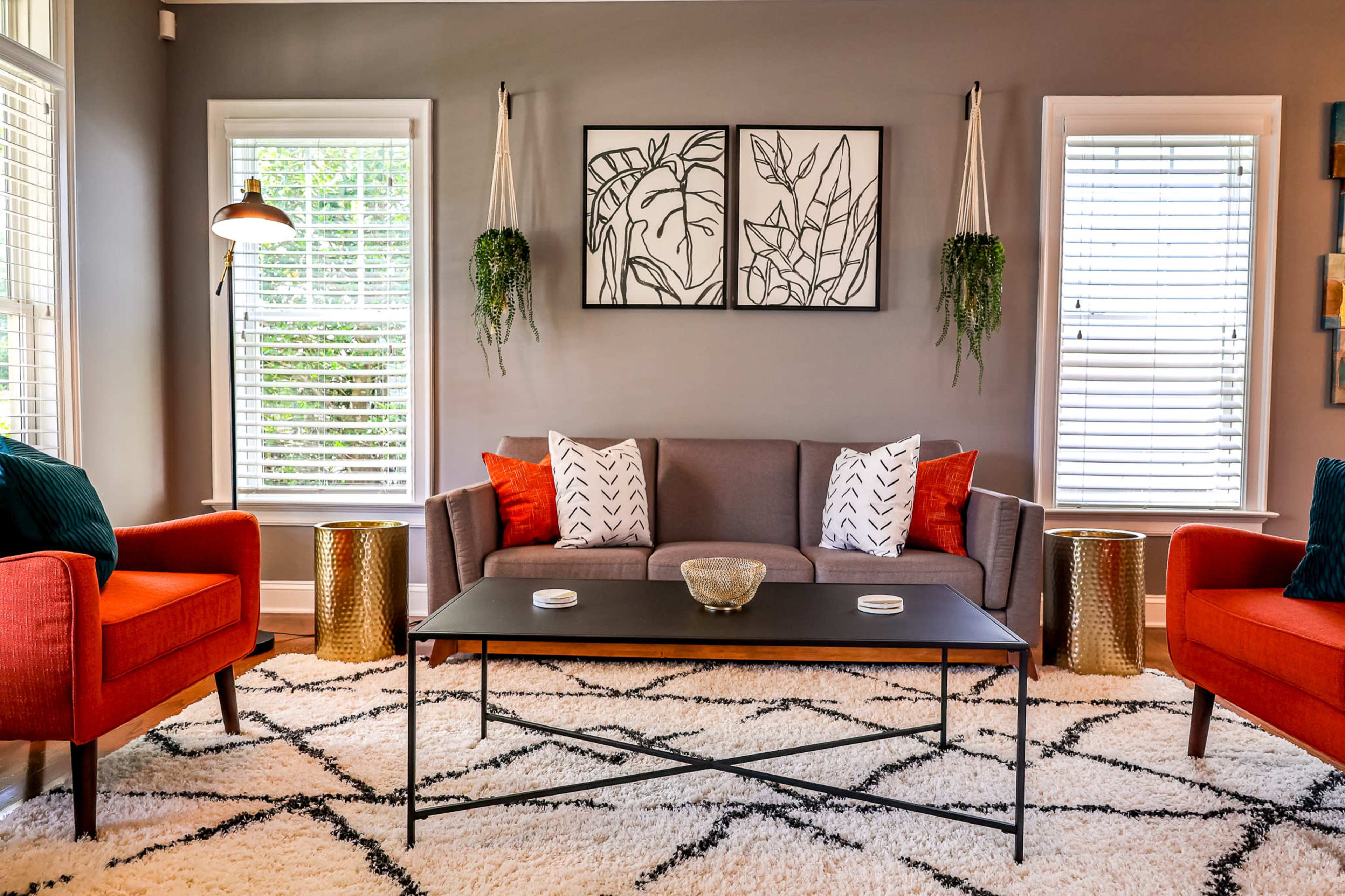 A living room features a gray sofa with colorful cushions, two red accent chairs, a black coffee table, and framed botanical prints on the wall.