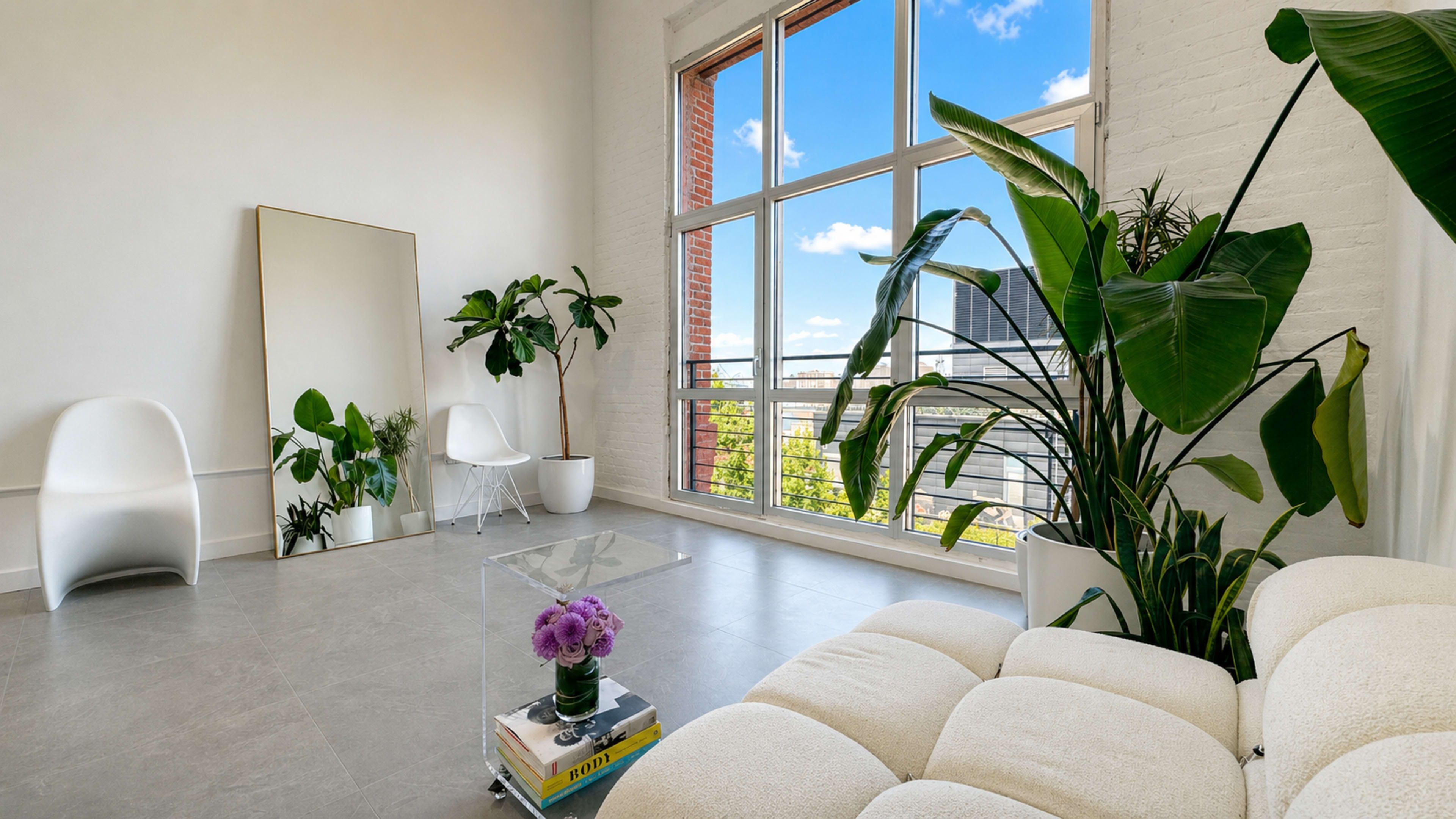 The image shows a bright, modern living room with large windows, a light-colored sofa, a glass coffee table, potted plants, and a decorative mirror.