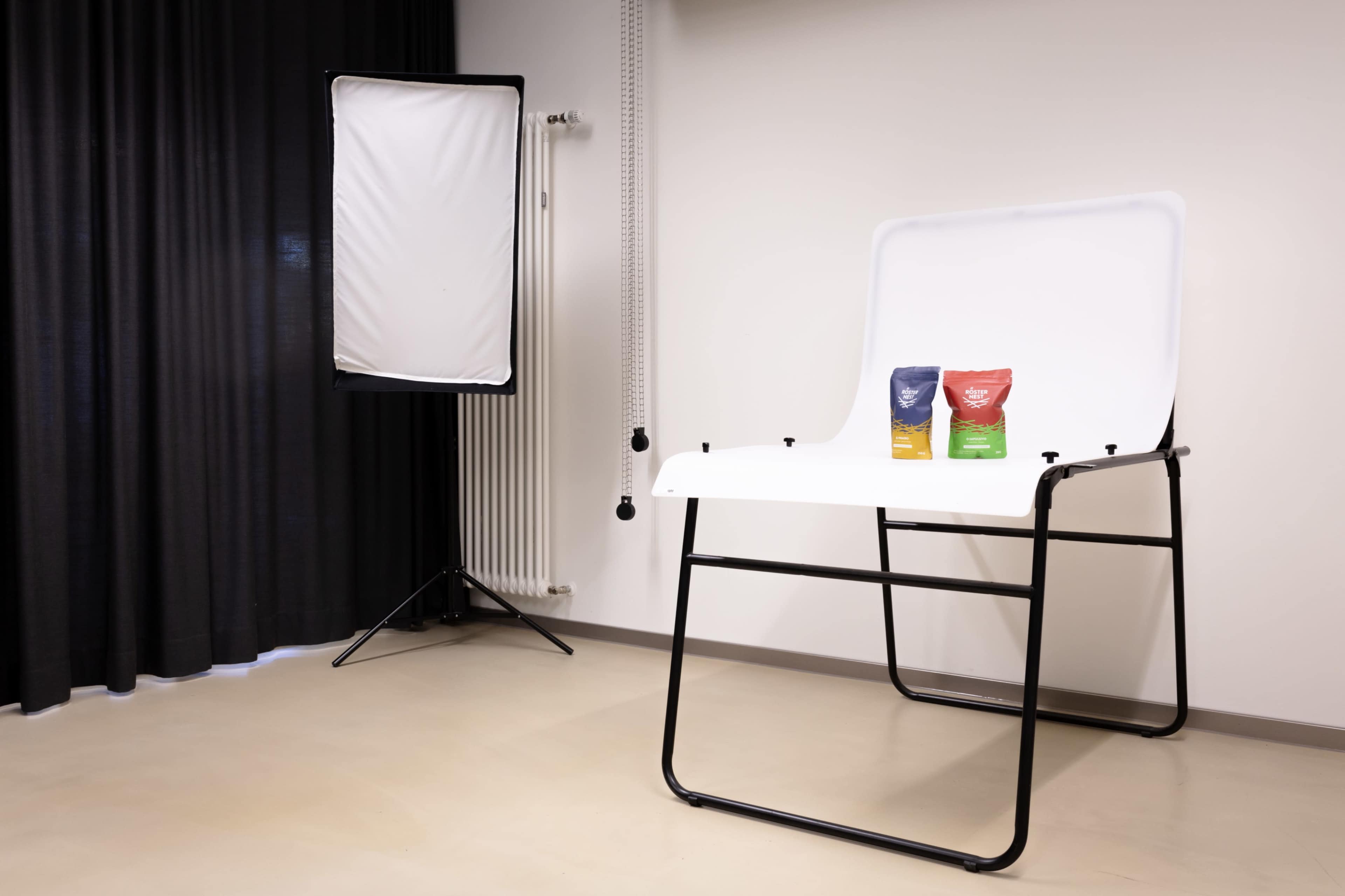 A photo studio setup features a white table with two bags of product placed on it, alongside a softbox lighting setup in a room with black curtains.