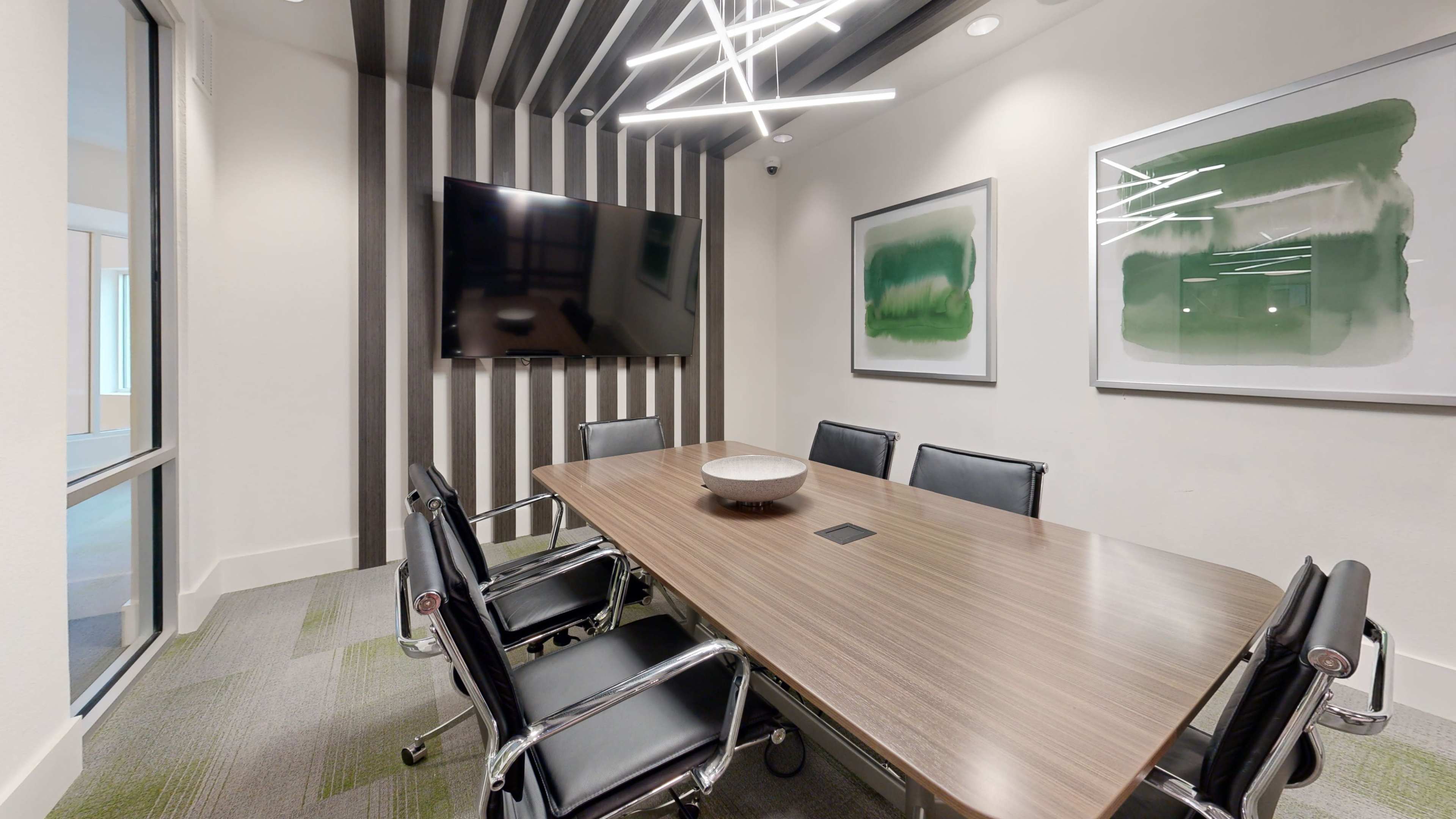A modern conference room features a long wooden table surrounded by black leather chairs, with a large television mounted on one wall and abstract green artwork on the adjacent walls.