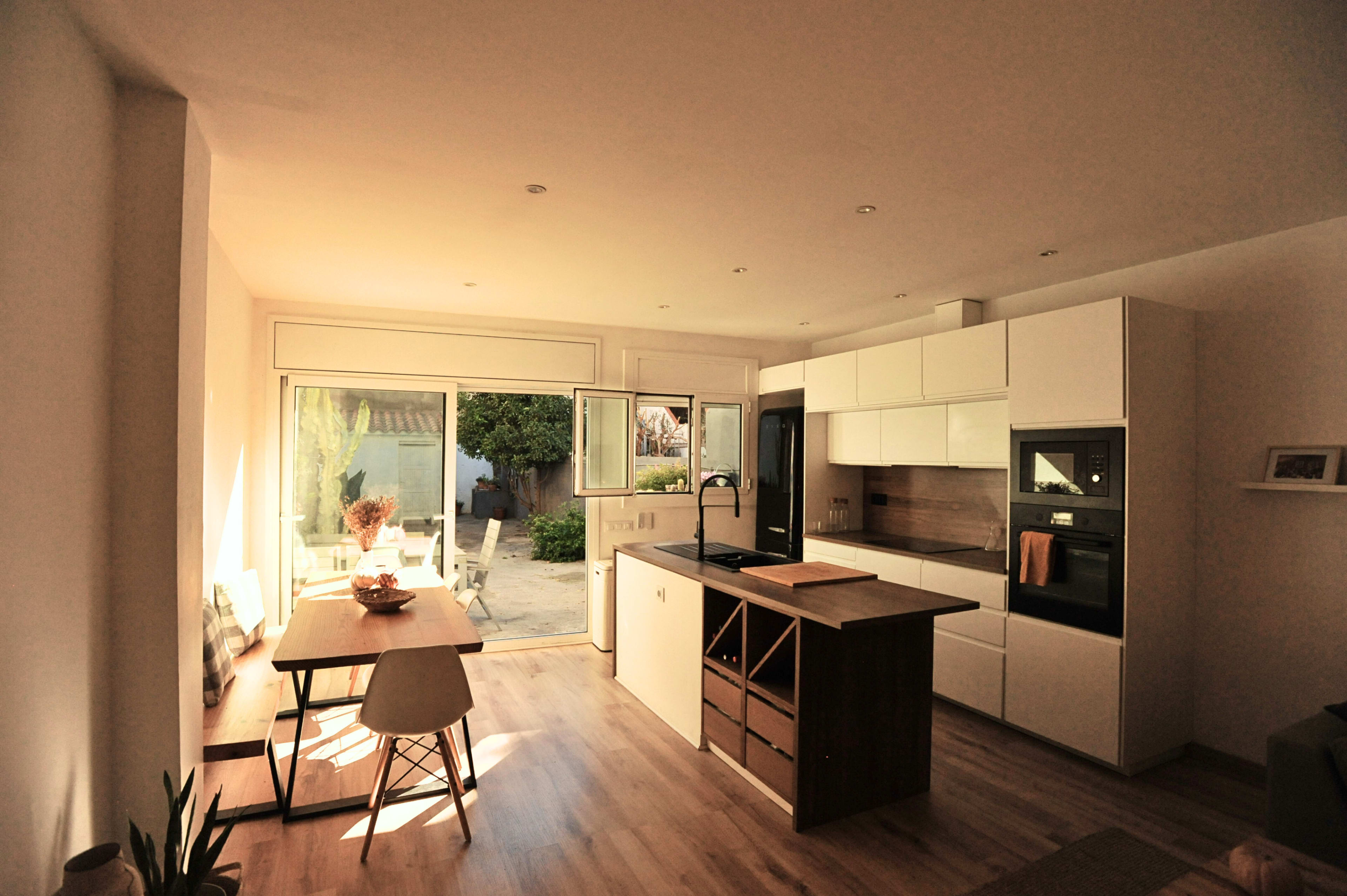 The image shows a modern kitchen-living area with a dining table, large windows allowing natural light, and minimalist cabinetry.