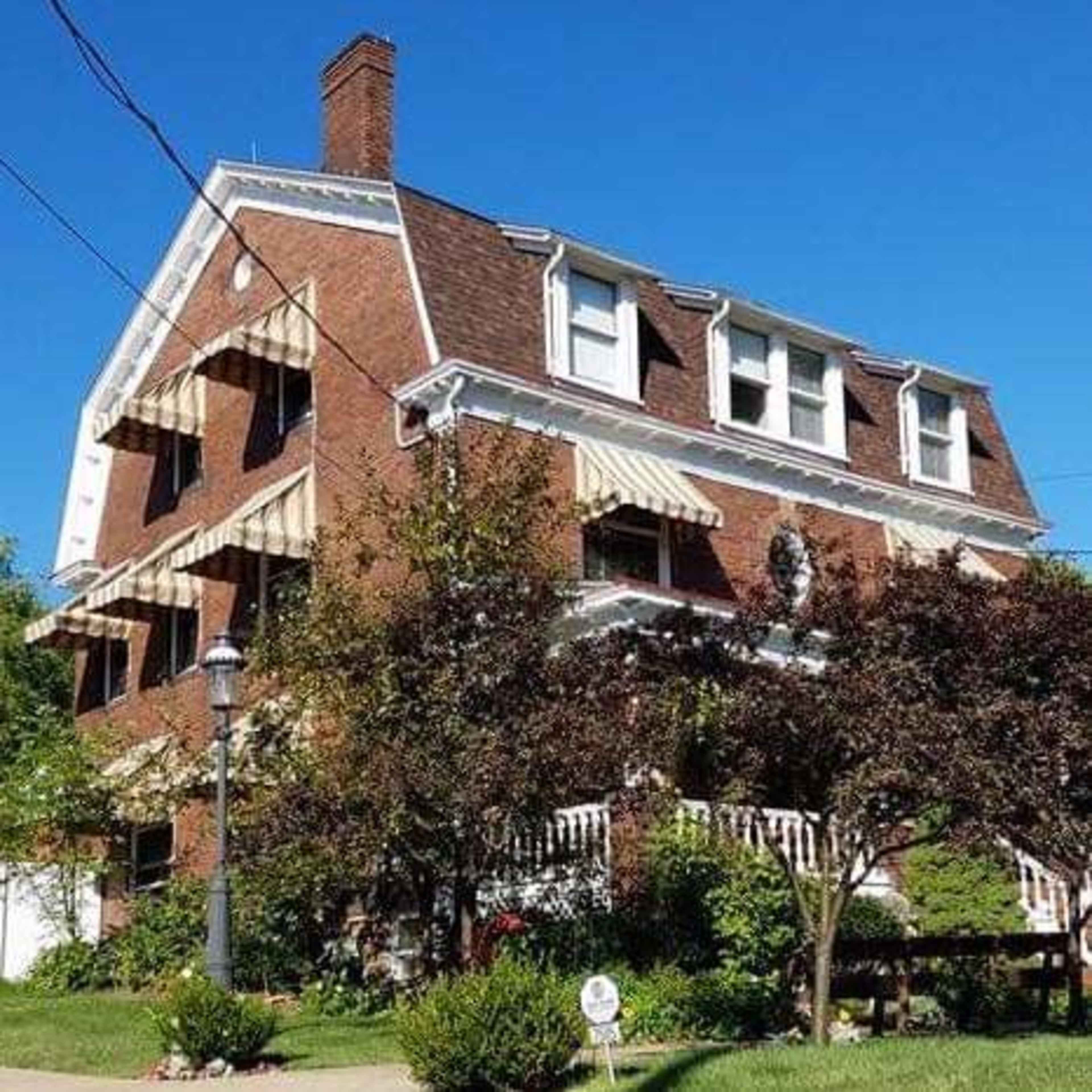 A three-story brick house features multiple awnings, a front porch, and landscaped greenery.