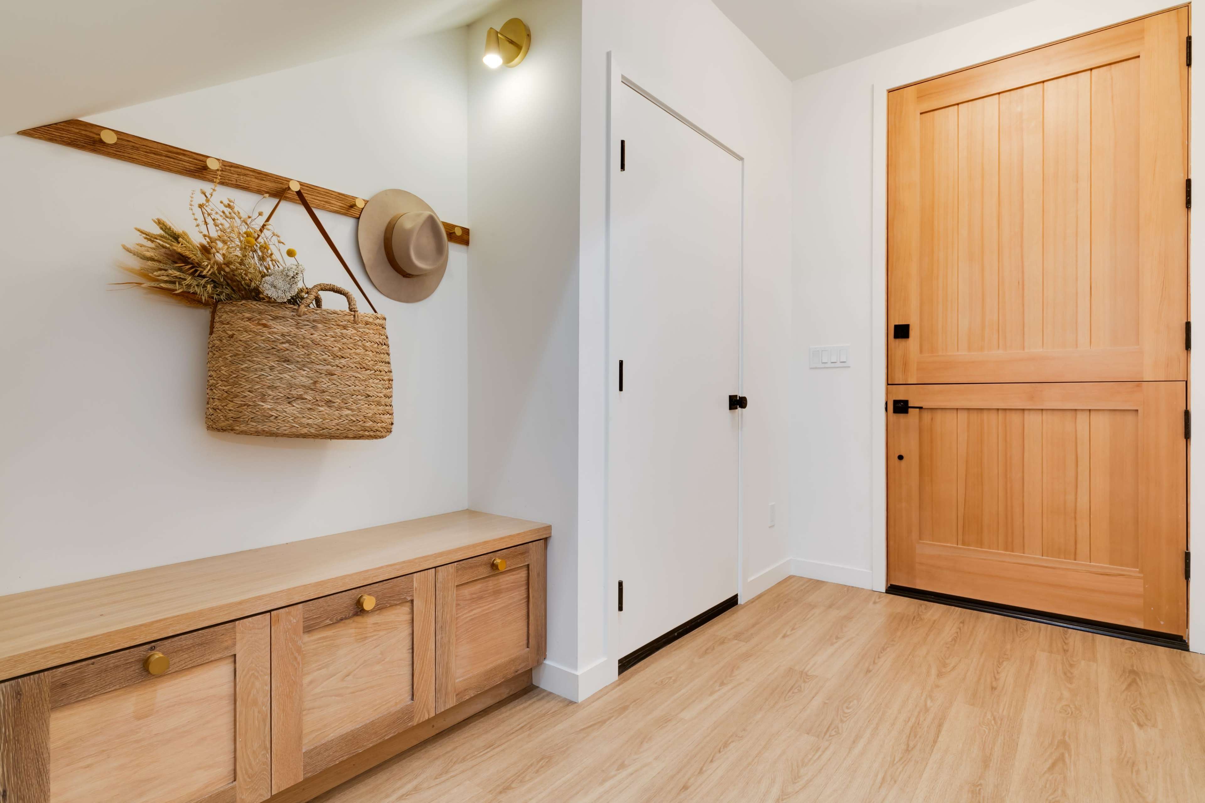 A modern entryway featuring a wooden bench, a door, a wall-mounted hat and basket holder, and light wood flooring.