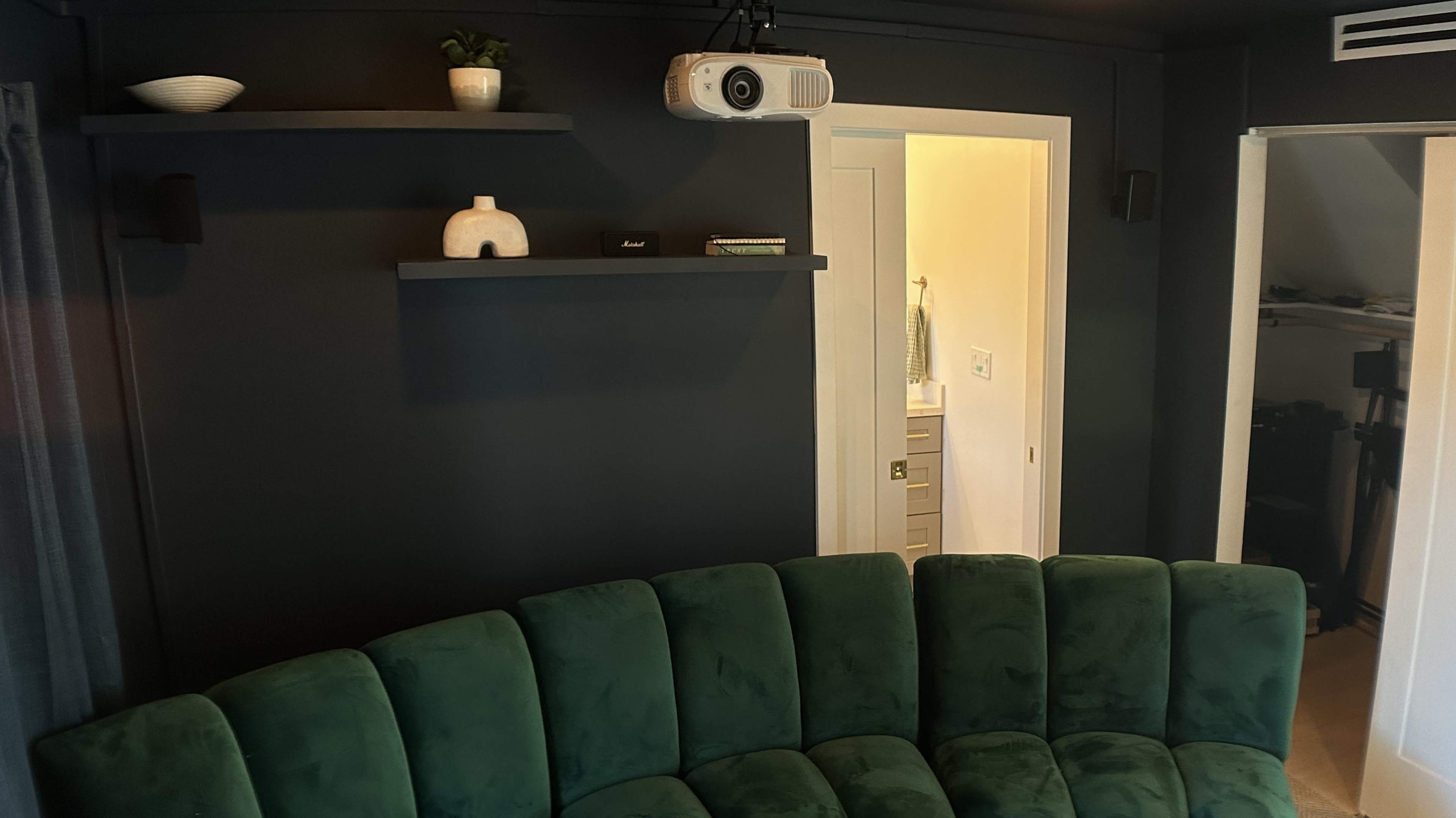 The image shows a cozy room with a green curved sofa, a projector mounted on the ceiling, and open doorways leading to adjacent spaces.