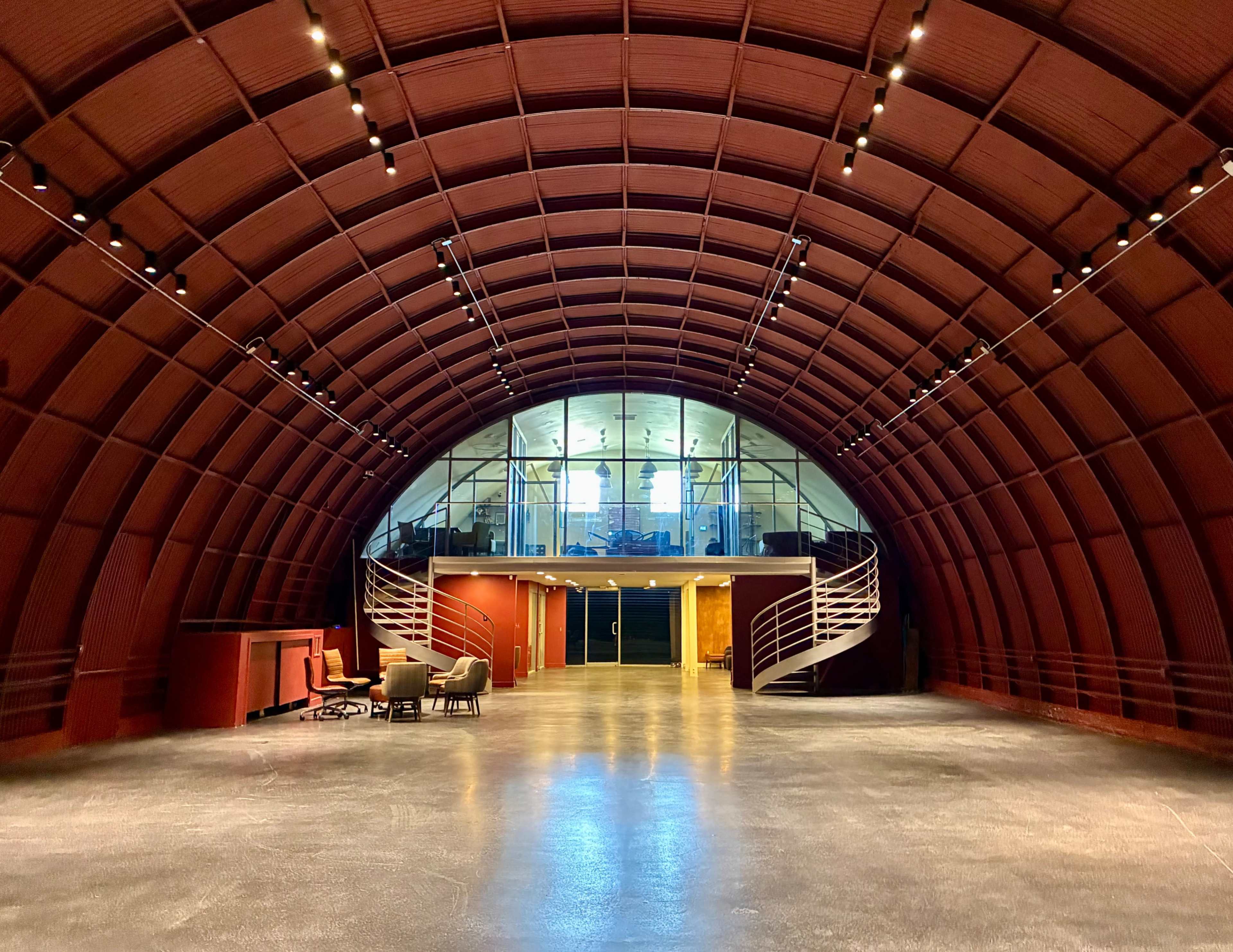 The image shows a spacious, modern interior with a large arched ceiling, featuring a polished concrete floor and a staircase leading to a glass-walled upper level.