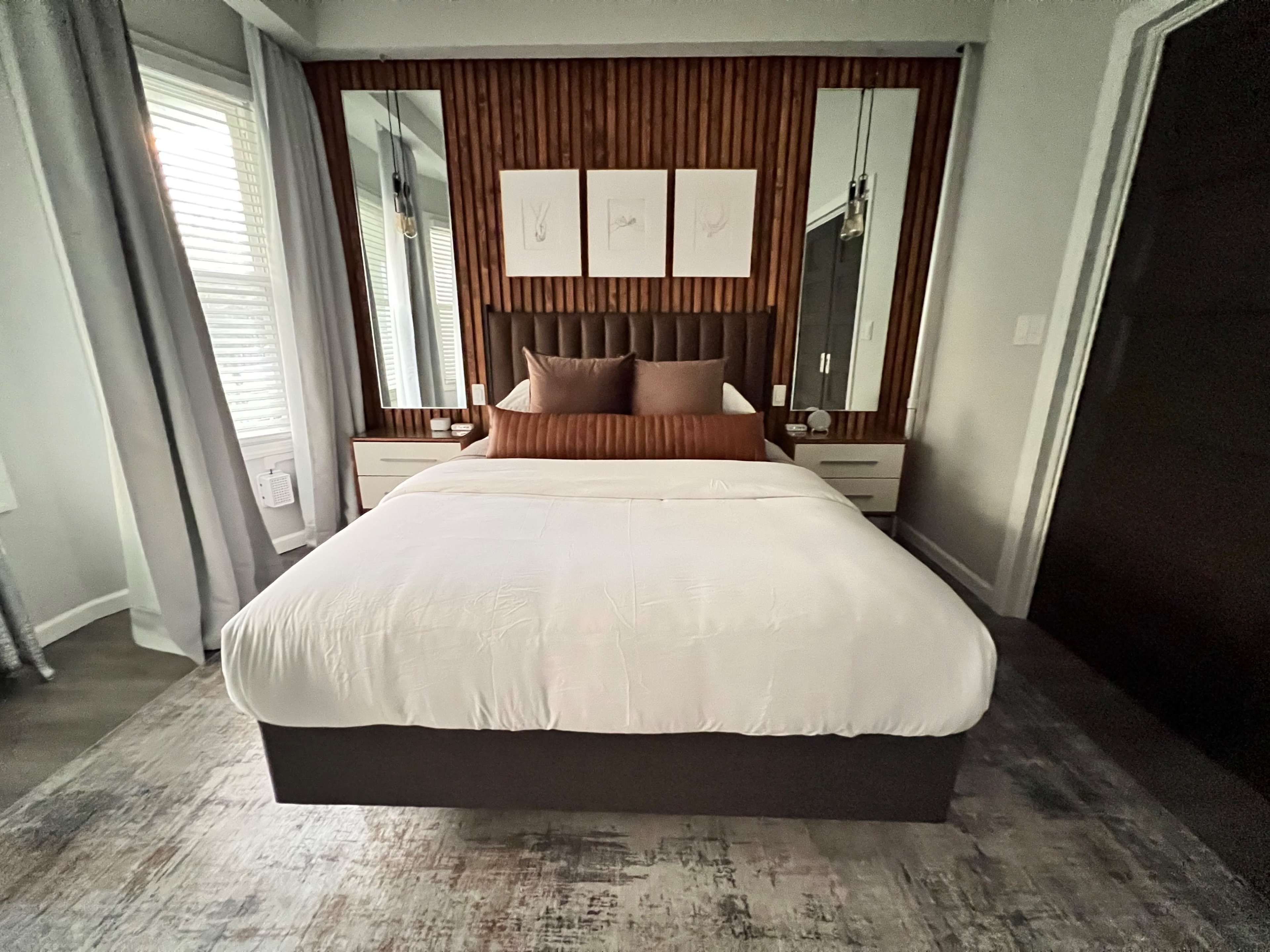 A modern bedroom featuring a queen-sized bed with a white comforter, brown accent pillows, and a mirrored headboard against a wall with wooden paneling.