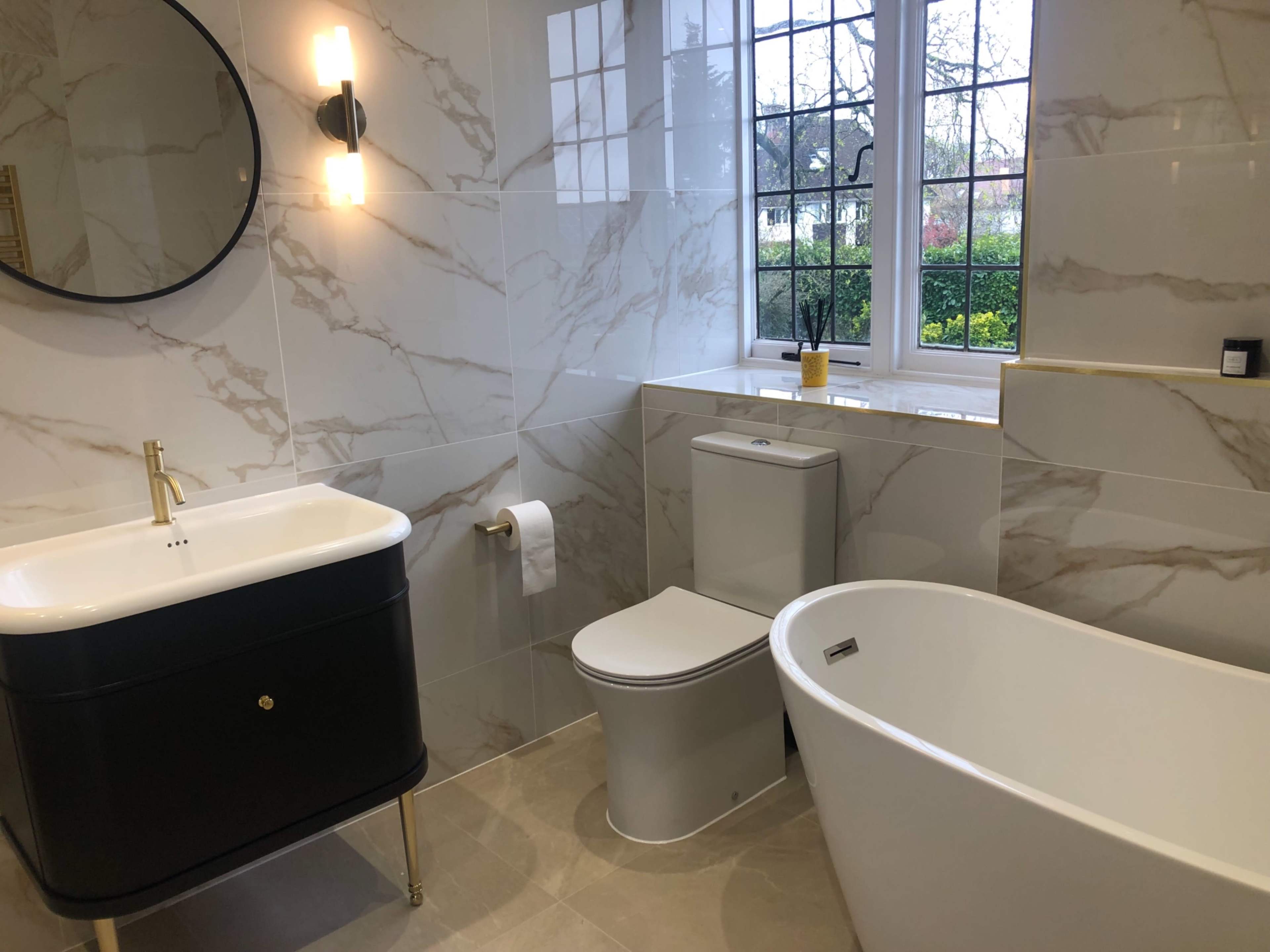 A modern bathroom featuring a freestanding bathtub, a toilet, and a vanity with a round mirror, all accented by marble-patterned walls and a window allowing natural light.