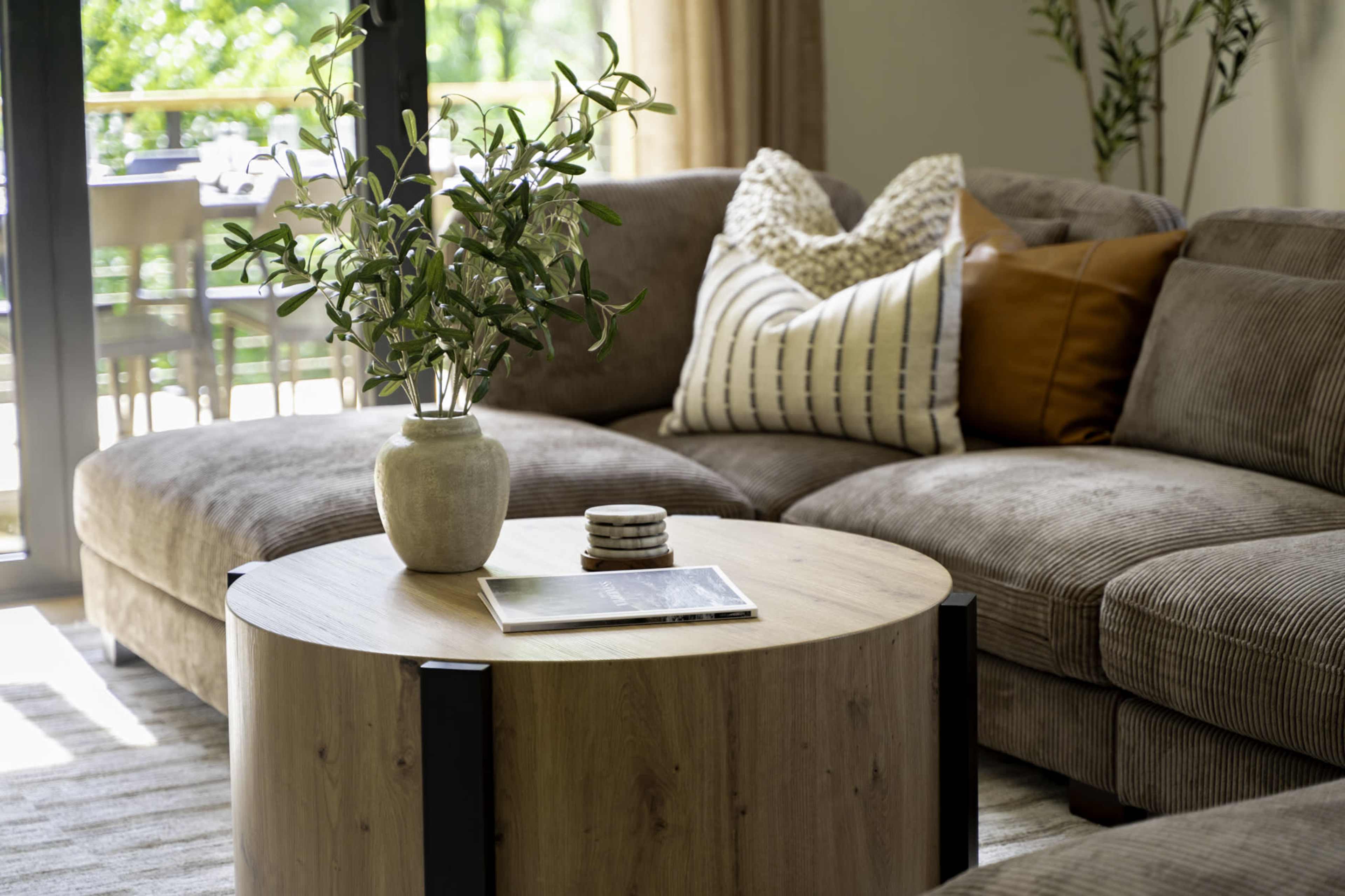 A spacious living room features a large, brown sectional sofa with decorative pillows and a round wooden coffee table topped with a vase of greenery and a magazine.